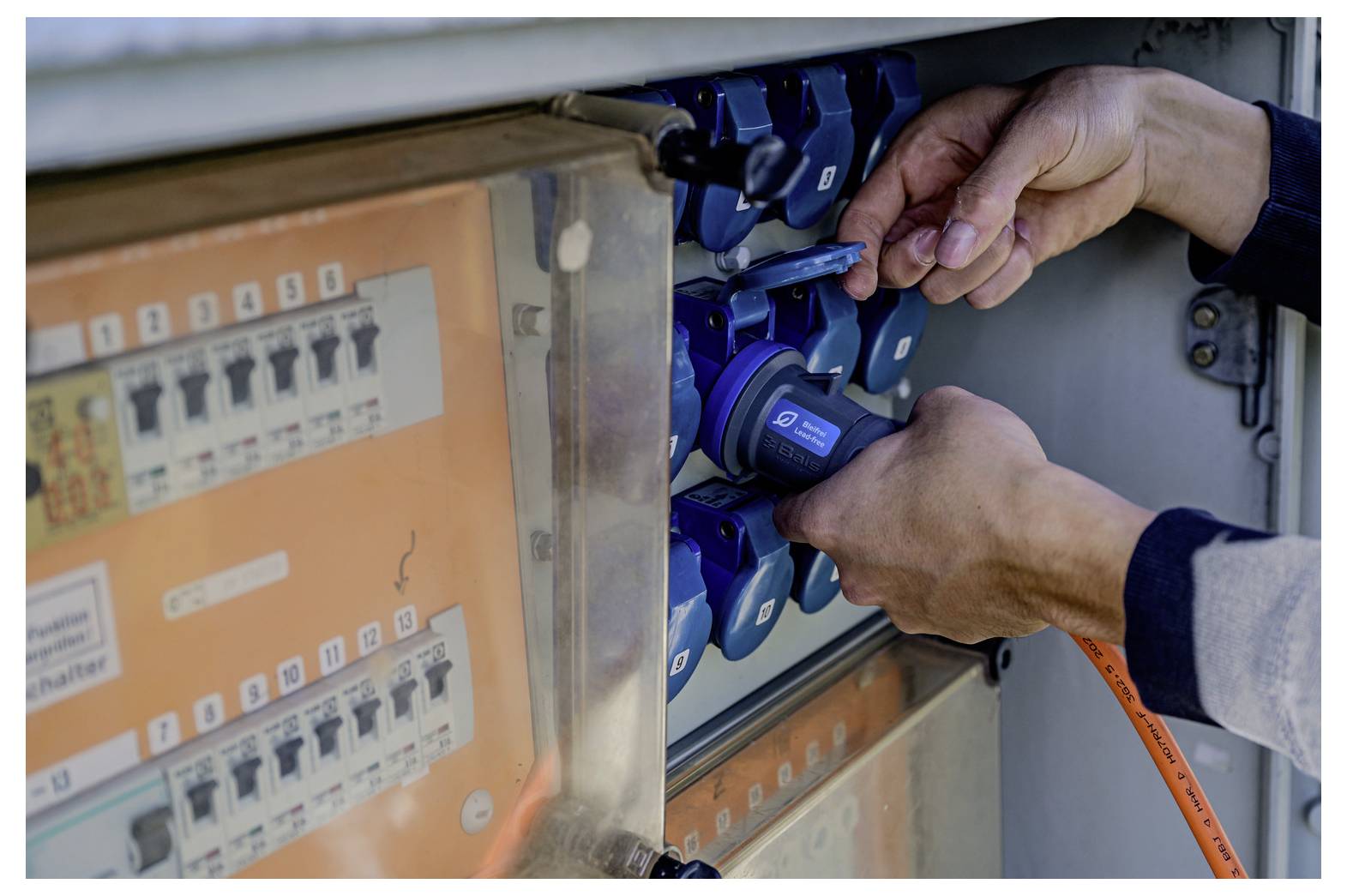 Hände stecken ein orangefarbenes Stromkabel in einen blauen Industriestromstecker an einer Schalttafel ein.