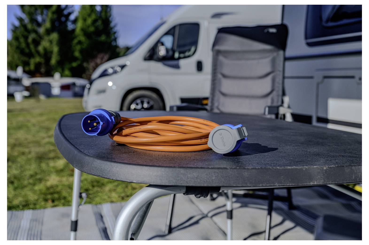 Oranges Stromkabel mit blauen und grauen Steckverbindern, das auf einem Campingtisch vor einem geparkten Wohnmobil aufgerollt ist
