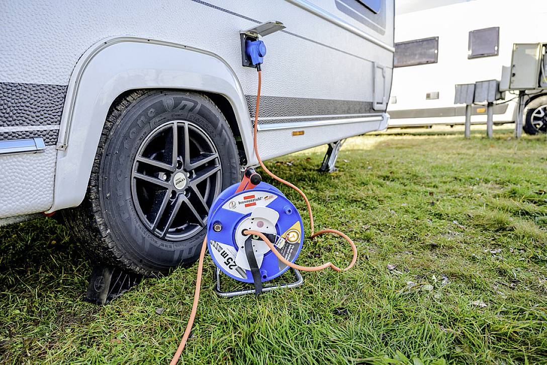 Wohnwagen mit angeschlossenem Verlängerungskabel zur Stromversorgung, geparkt auf Rasenfläche. Kabeltrommel liegt am Boden.