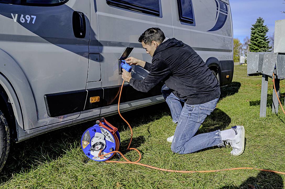 Eine Person kniet auf einer Wiese und schließt ein Verlängerungskabel an ein Wohnmobil an. Im Hintergrund sind Bäume sichtbar.