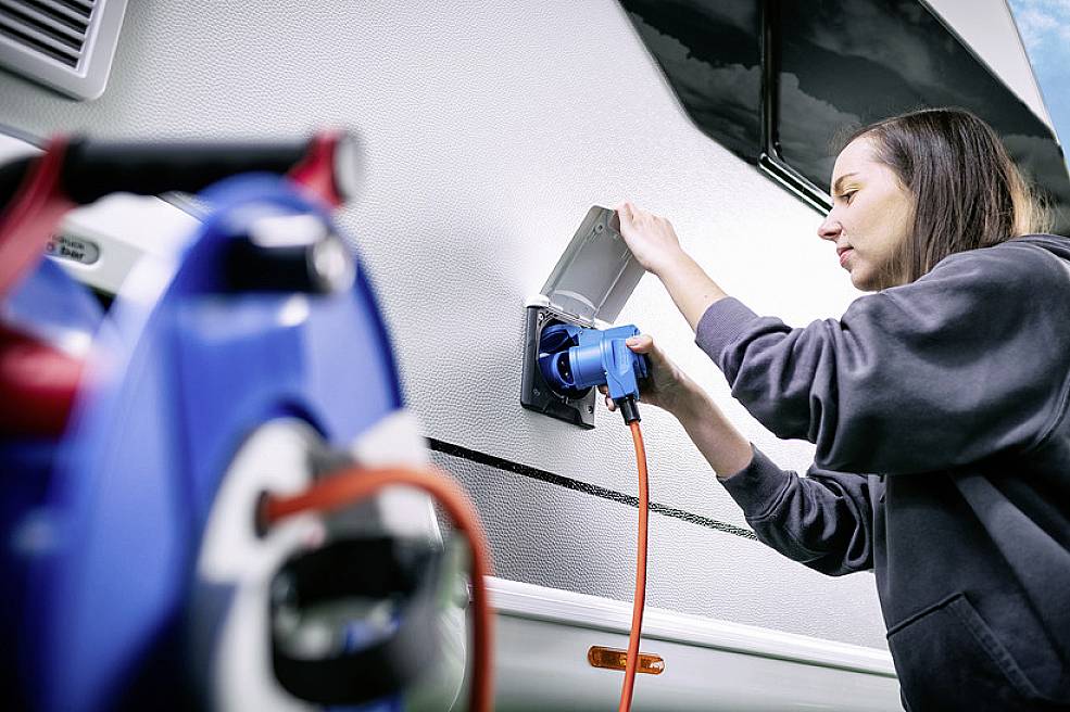 Eine Frau schließt ein Elektrokabel an ein Wohnmobil an, um es aufzuladen. Im Vordergrund ist ein blauer Stecker erkennbar.