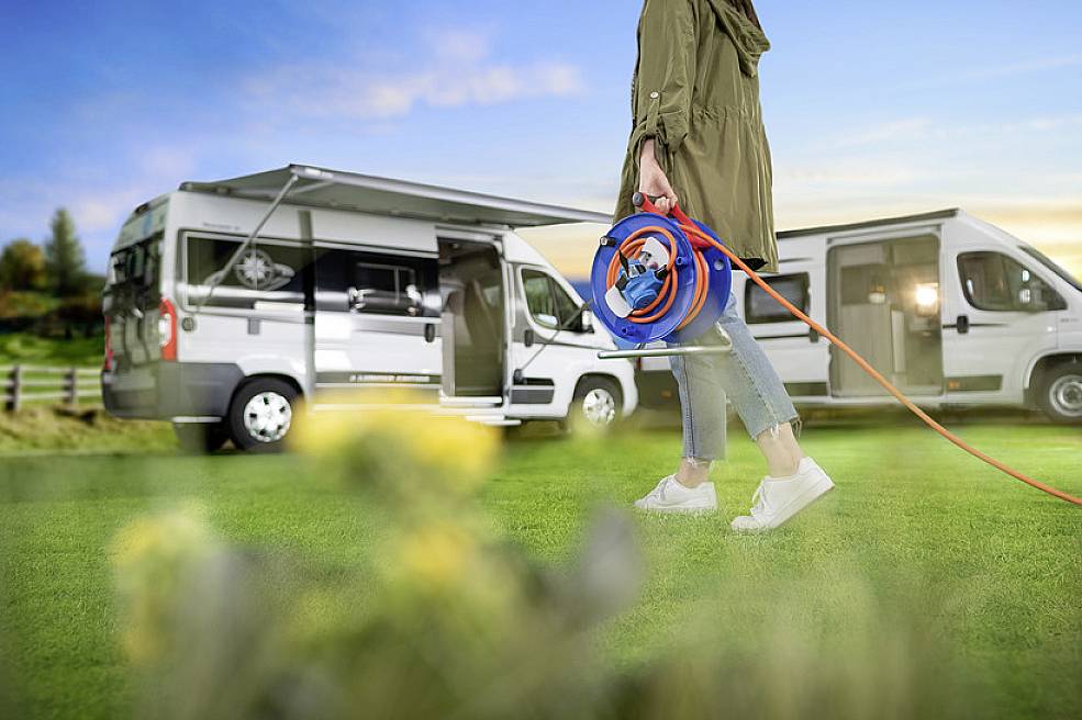 Eine Person in Freizeitkleidung hält eine Kabeltrommel vor einem geparkten Wohnmobil mit offenem Türbereich in einer grünen Landschaft.