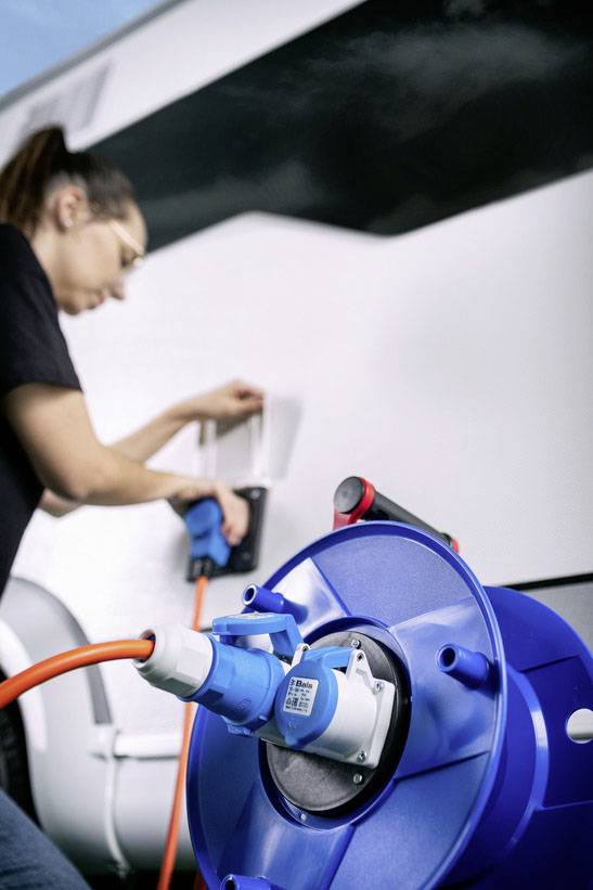 Eine Person steckt ein Elektrokabel in ein Wohnmobil, das mit einer blauen Stromtrommel verbunden ist.