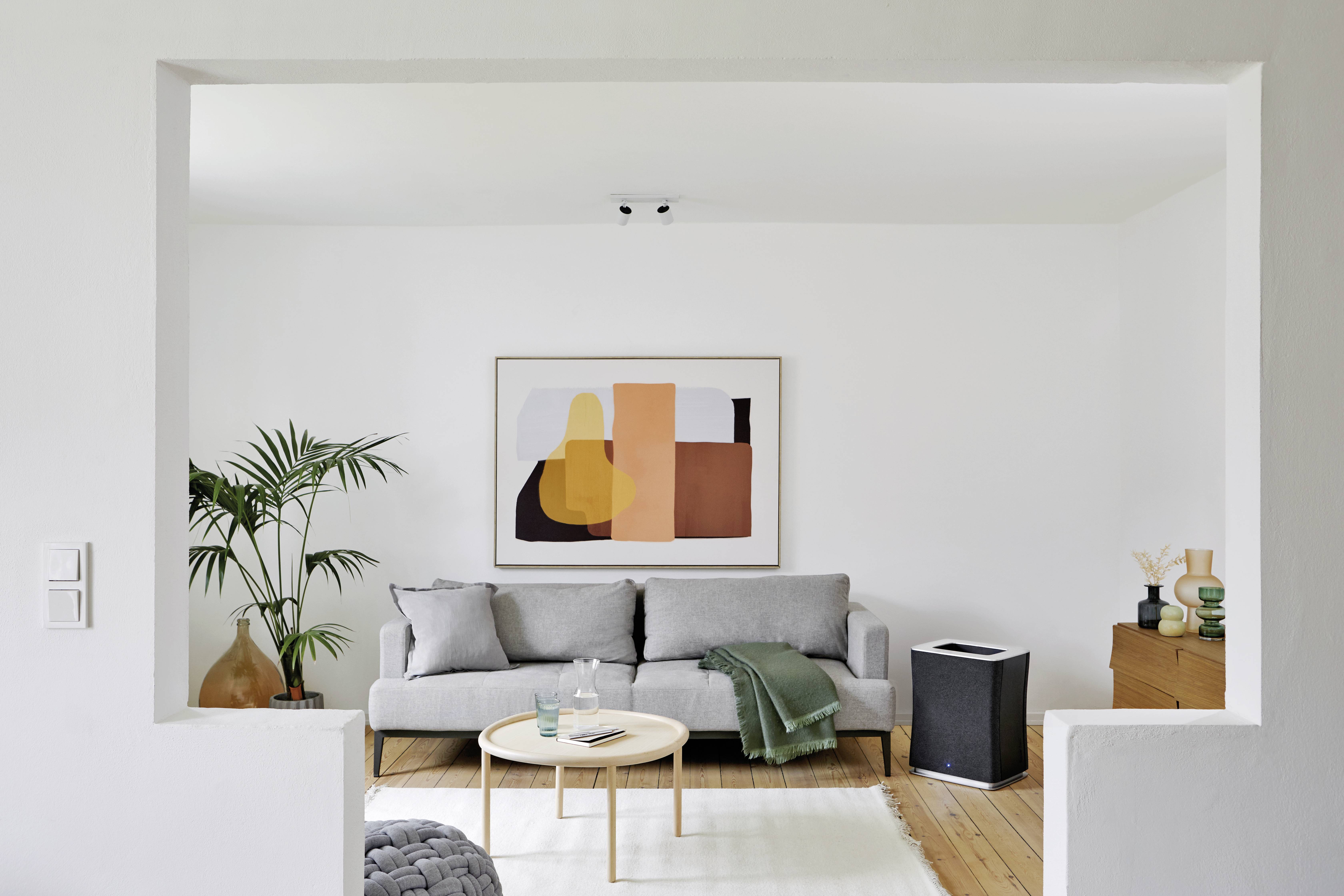 Ein modernes Wohnzimmer mit grauem Sofa, großem Pflanzengefäß, rundem Holztisch und abstrakter Kunst an der weißen Wand.