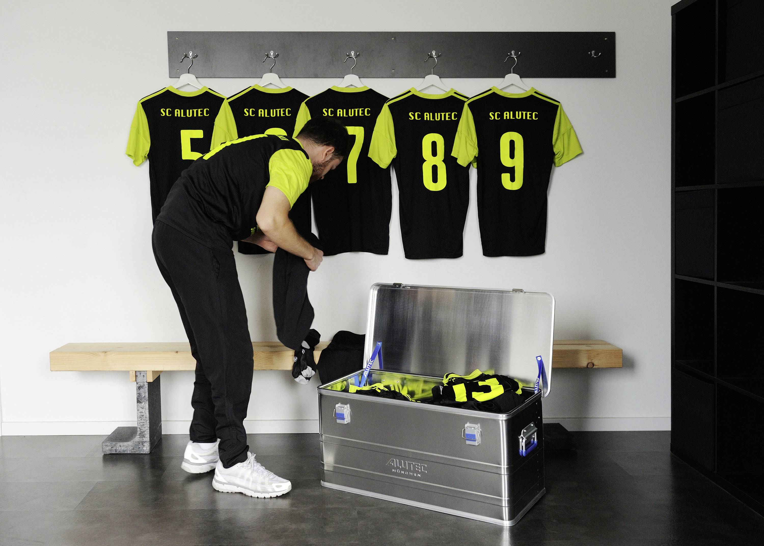 Ein Mann im Umkleideraum packt Sportkleidung aus einer Metallkiste aus. Vier Trikots mit gelben Nummern hängen an der Wand.