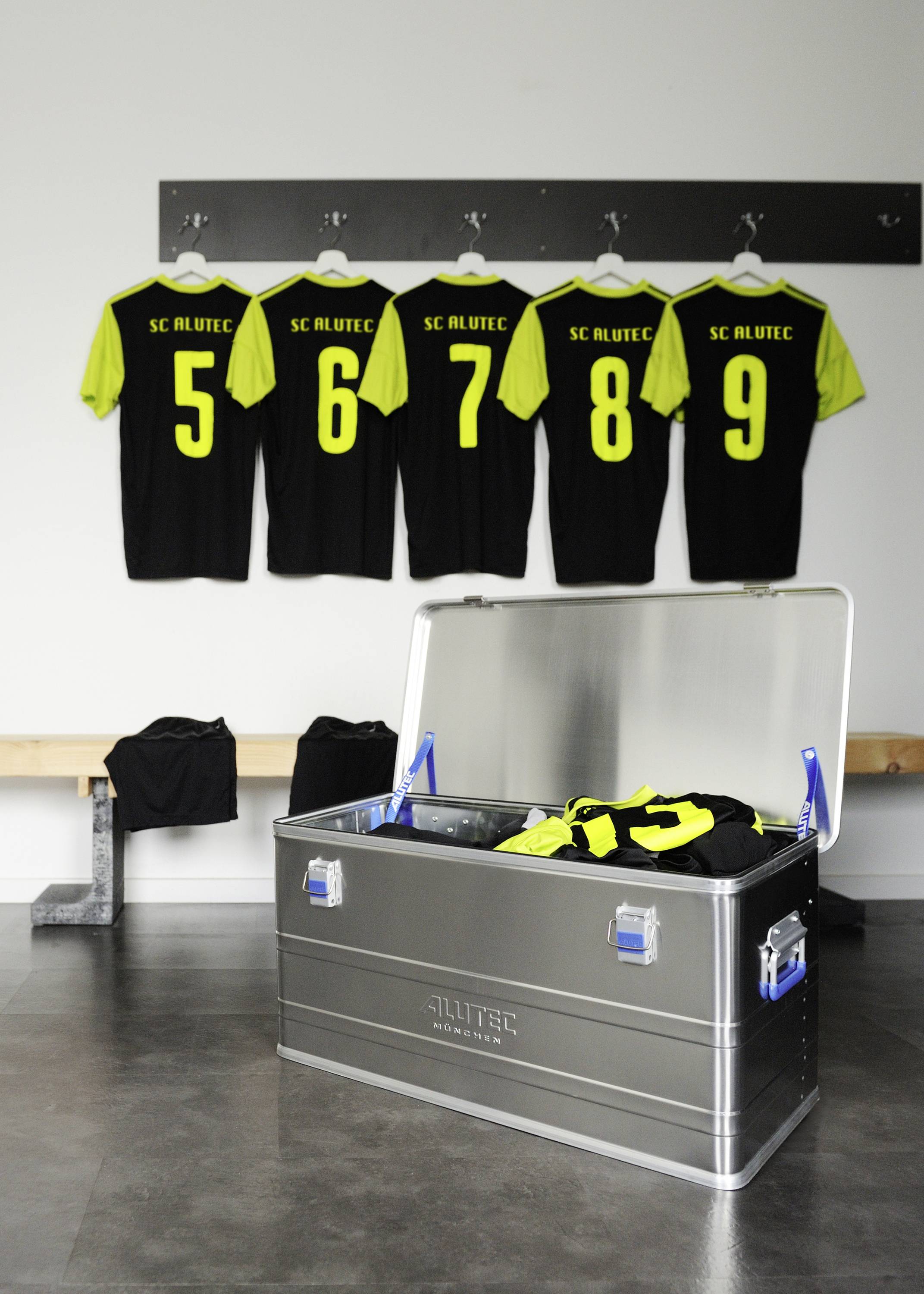 Fünf Fußballtrikots mit den Nummern 5 bis 9 hängen an der Wand. Eine geöffnete Metallkiste mit anderen Trikots steht im Vordergrund.