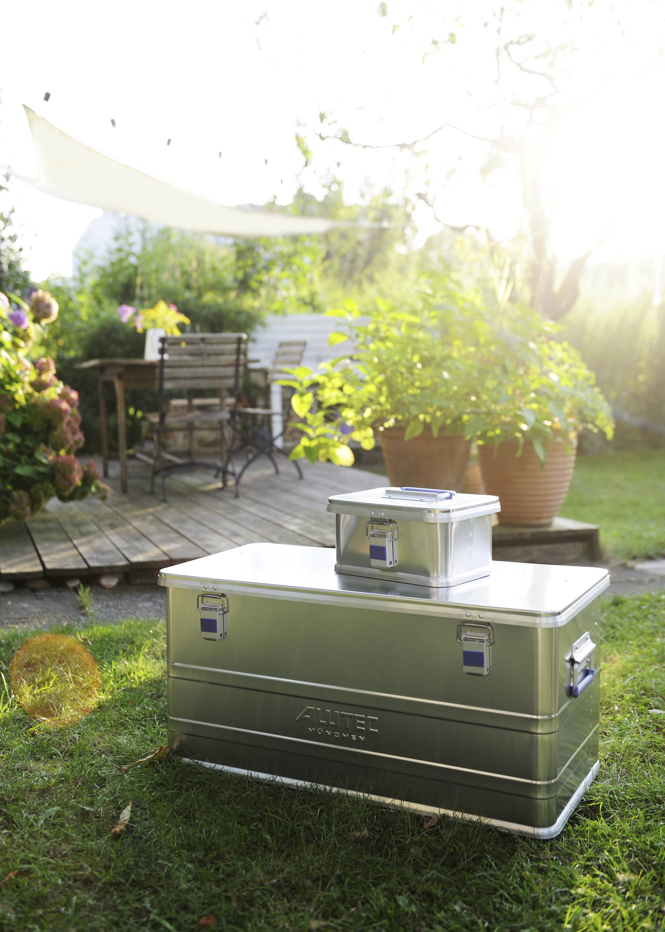 Zwei Metallkisten stehen auf einer Wiese im Garten, im Hintergrund ein Tisch mit Stühlen auf einer Holzterrasse, sonniges Wetter.