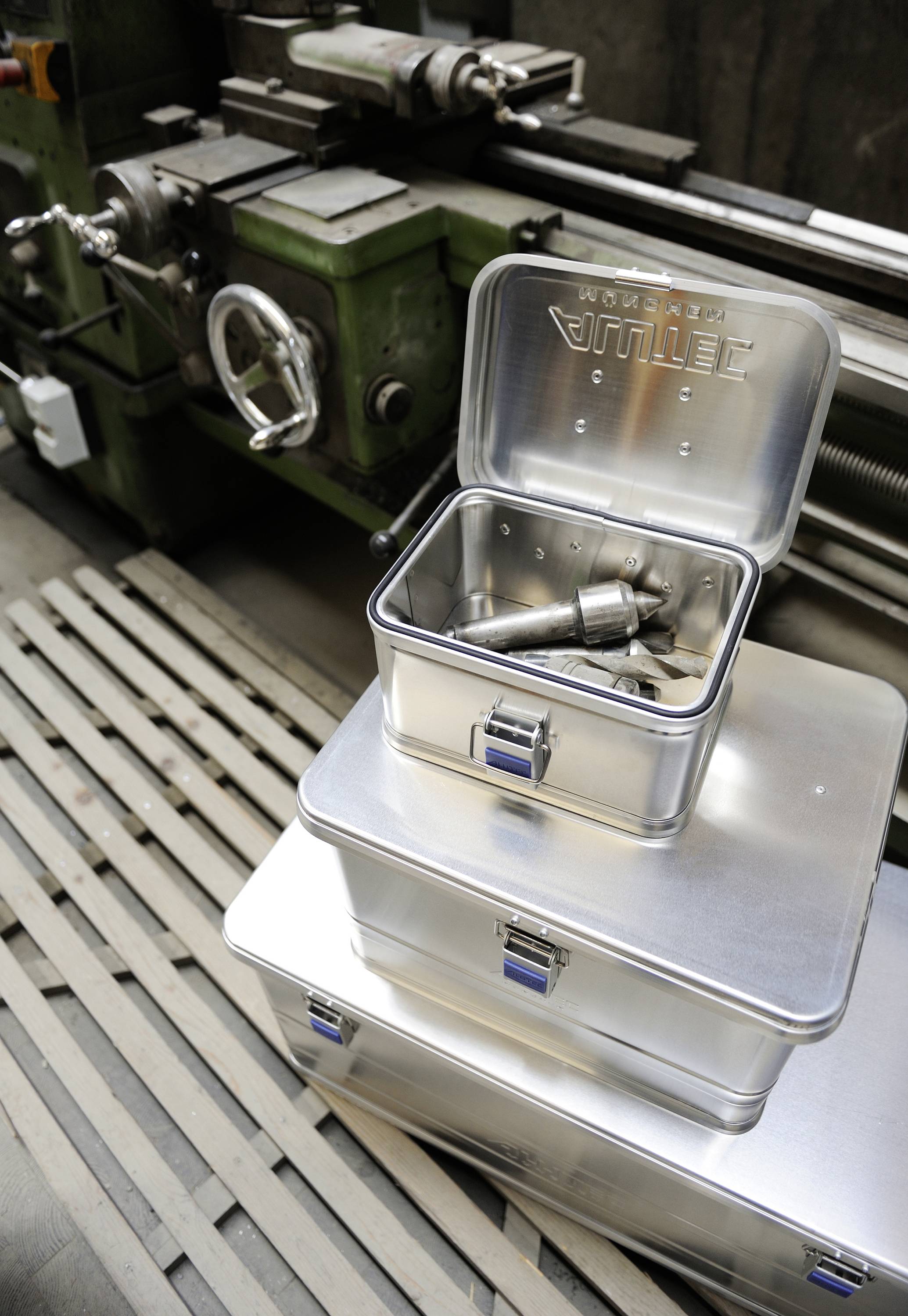 Drei Metallboxen im Vordergrund in einer Werkstatt; eine offen mit Werkzeugen. Im Hintergrund eine grüne Drehmaschine.