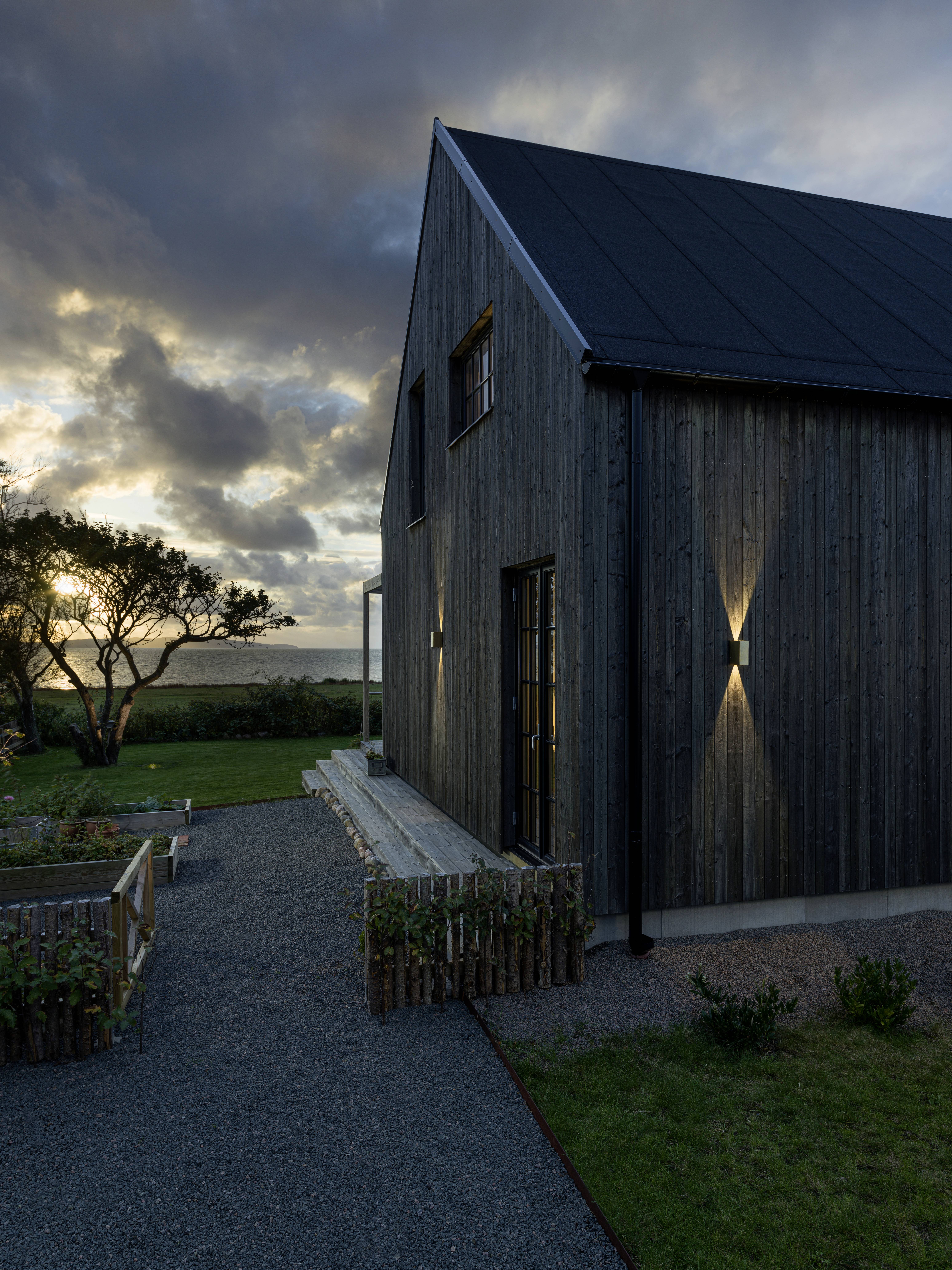 Ein modernes, hölzernes Haus bei Sonnenuntergang mit einem Garten und Bäumen im Hintergrund, unter wolkigem Himmel.