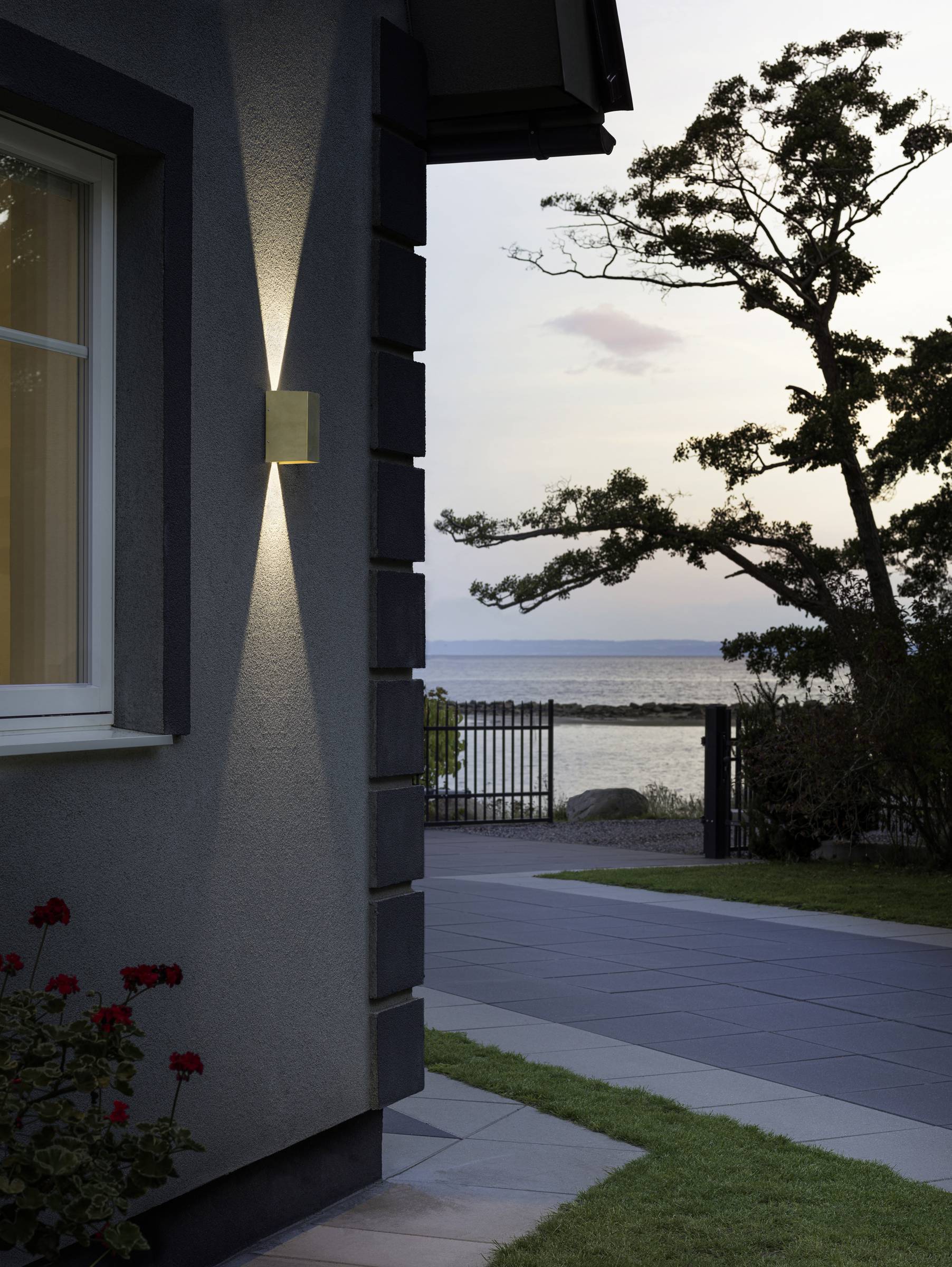 Hausfassade bei Sonnenuntergang, Außenlampe leuchtet, Fenster zeigt Gardine. Baum und Meer im Hintergrund, ruhige Atmosphäre.