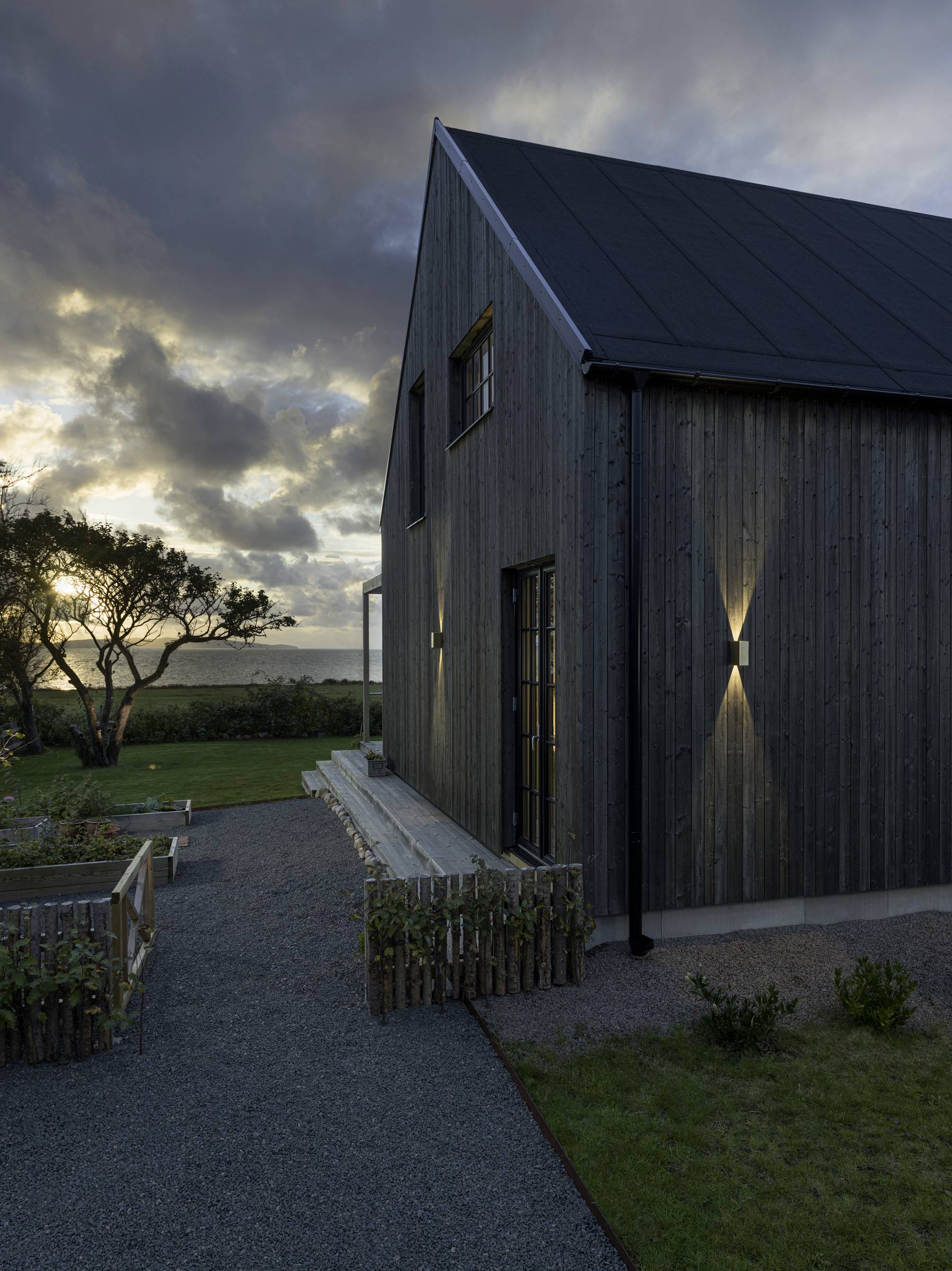 Ein modernes Holzhaus bei Sonnenuntergang. Der Himmel ist bewölkt, und im Hintergrund sieht man Bäume und eine Wiese.
