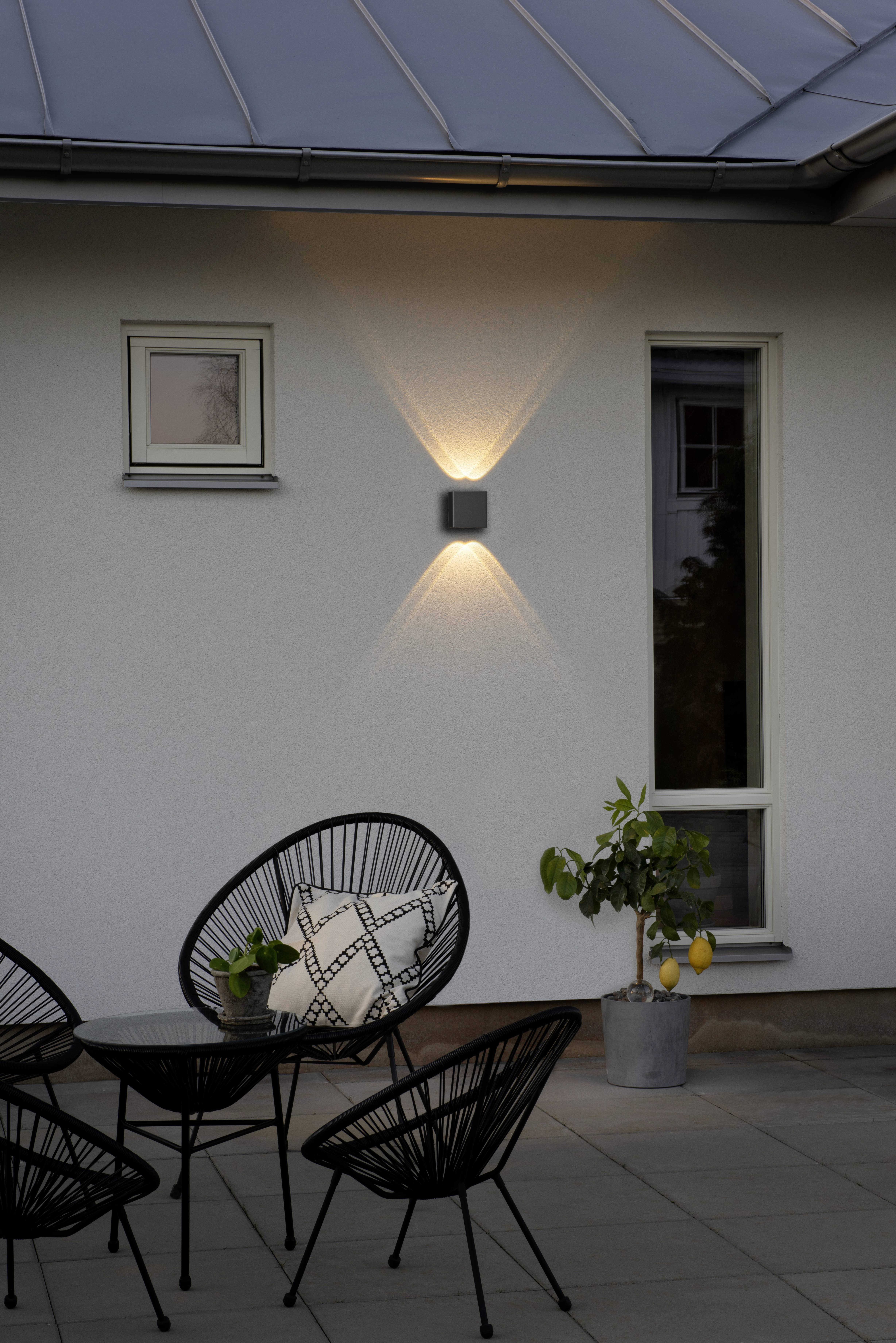 Eine Außenwand mit einer modernen Wandleuchte, darunter ein Tisch mit schwarzen Rattanstühlen. Rechts steht ein kleiner Zitronenbaum.