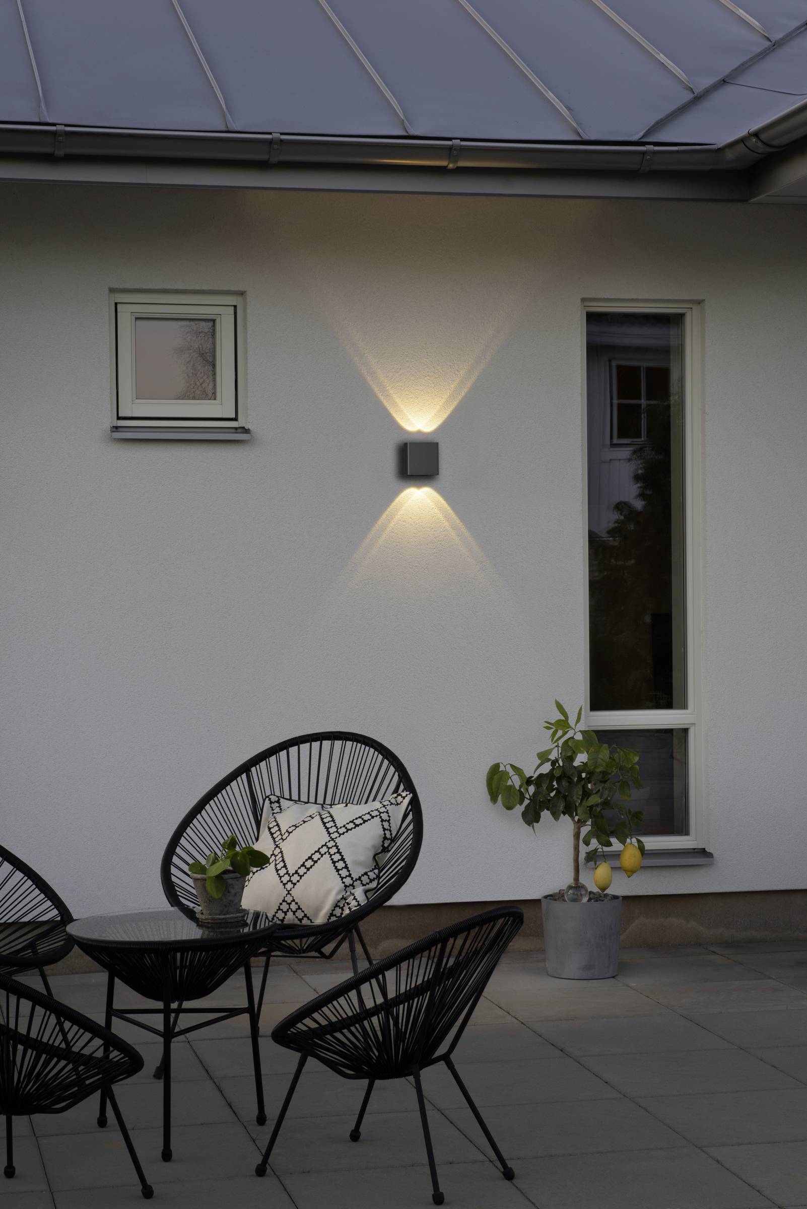 Moderne Terrasse mit anthrazitfarbenen Stühlen und Tisch, Wandlampe leuchtet, kleines Zitronenbäumchen daneben, Fenster im Hintergrund.