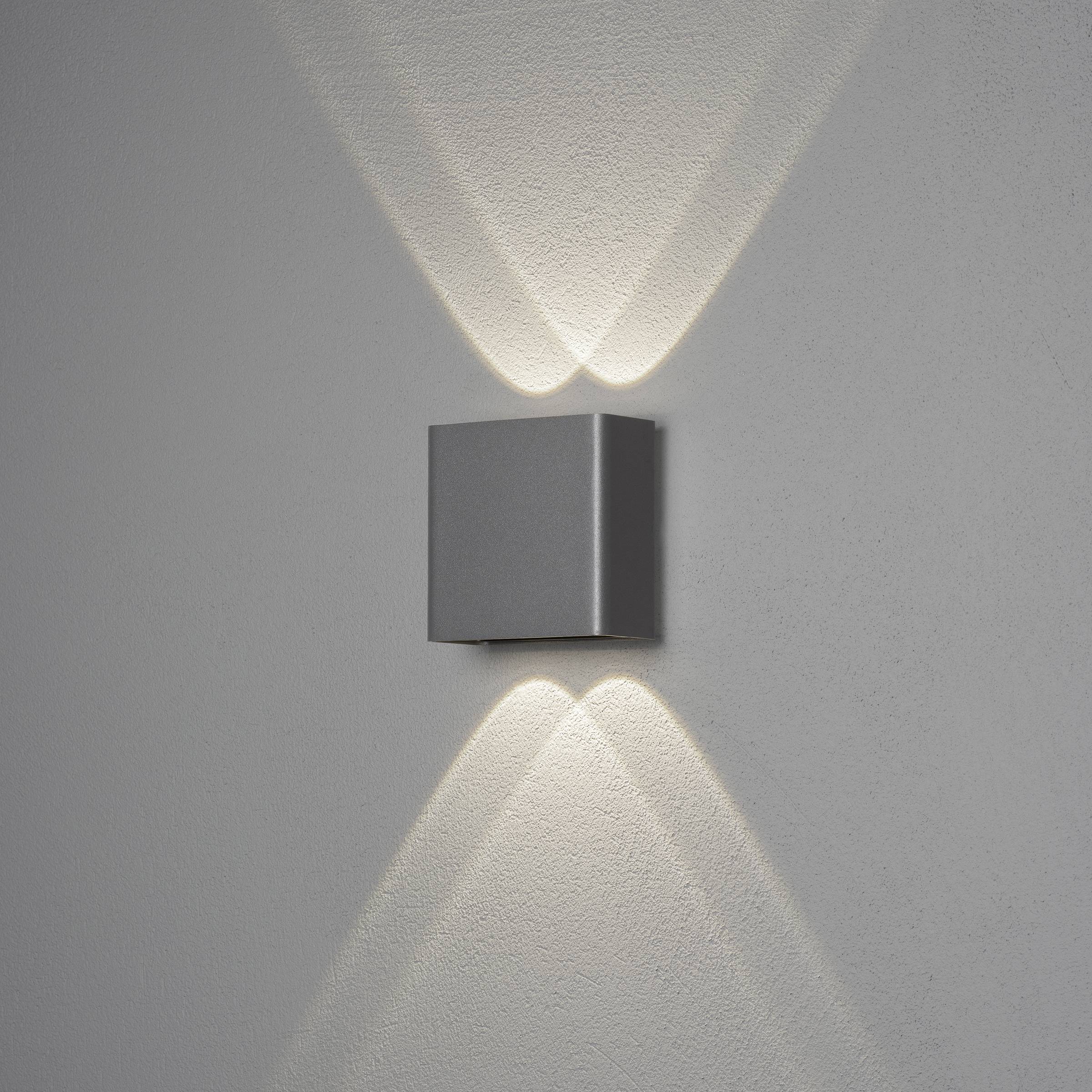 Silberne Wandleuchte mit würfelförmigem Gehäuse, strahlt sanftes Licht nach oben und unten, erzeugt ein symmetrisches Muster.