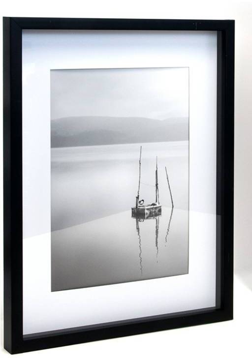 Ein Schwarzweißfoto zeigt ein ruhiges Seeufer mit Bergen im Hintergrund und ein kleines Boot mit Segeln im Vordergrund.