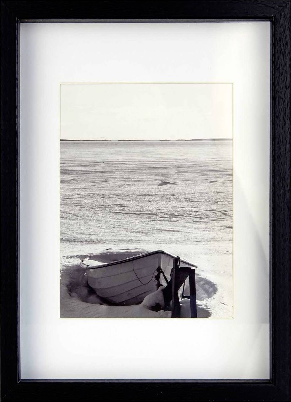 Ein altes Boot liegt verlassen im Schnee am Ufer eines gefrorenen Sees unter einem bewölkten Himmel, eingefasst in einem schwarzen Rahmen.
