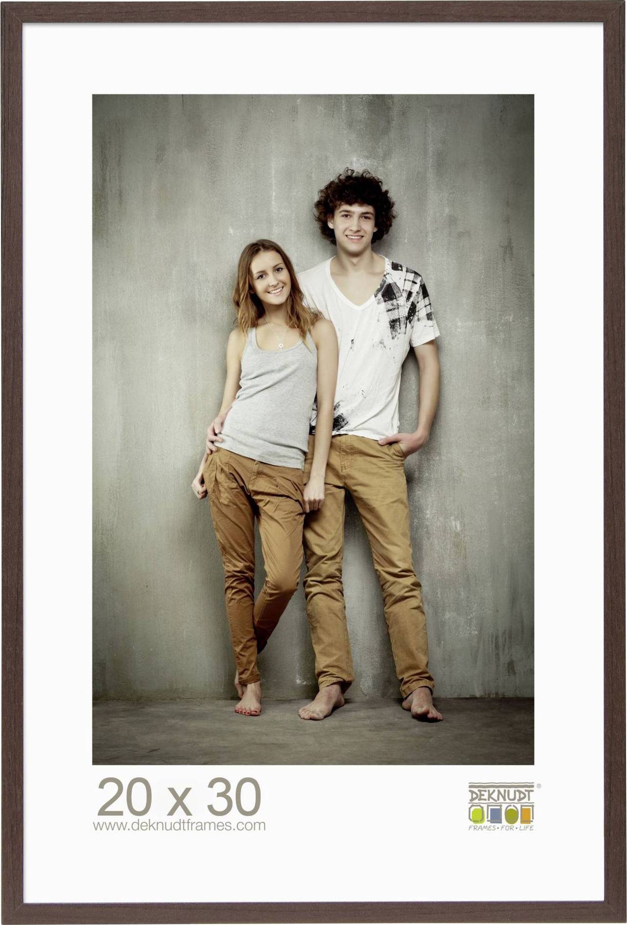Ein lächelndes Paar posiert barfuß vor einer grauen Wand, beide tragen beige Hosen. Darunter steht '20 x 30 www.deknudtframes.com'.