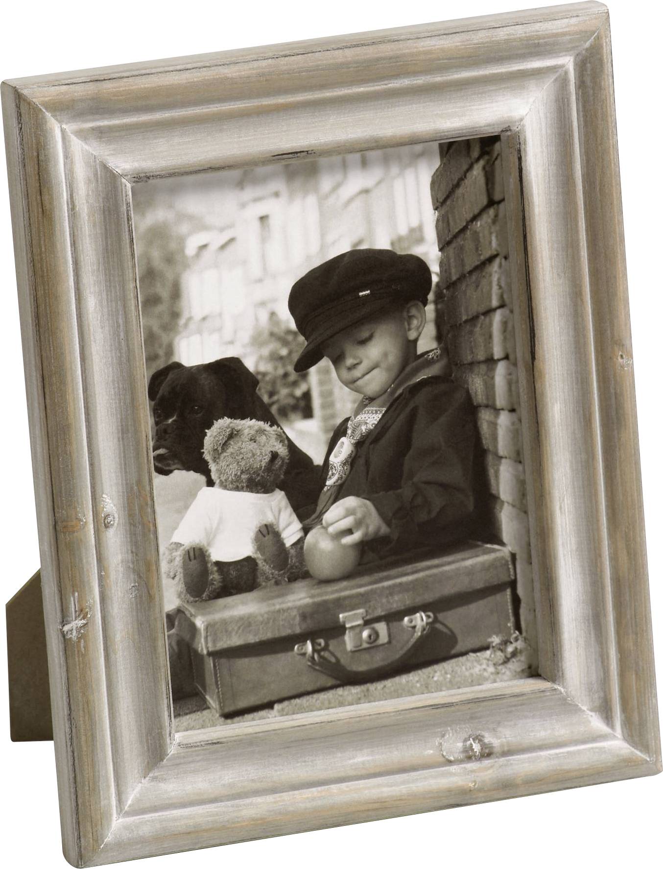 Ein Kind mit Mütze und Hund sitzt an einer Backsteinmauer, hält einen Apfel und sitzt neben einem Teddybär auf einem Koffer.