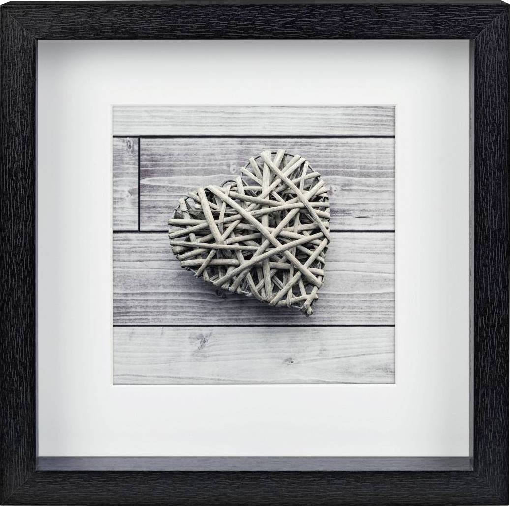 Ein herzförmiges Geflecht aus Zweigen liegt auf einer Holzoberfläche und ist in einem schwarzen Rahmen präsentiert.