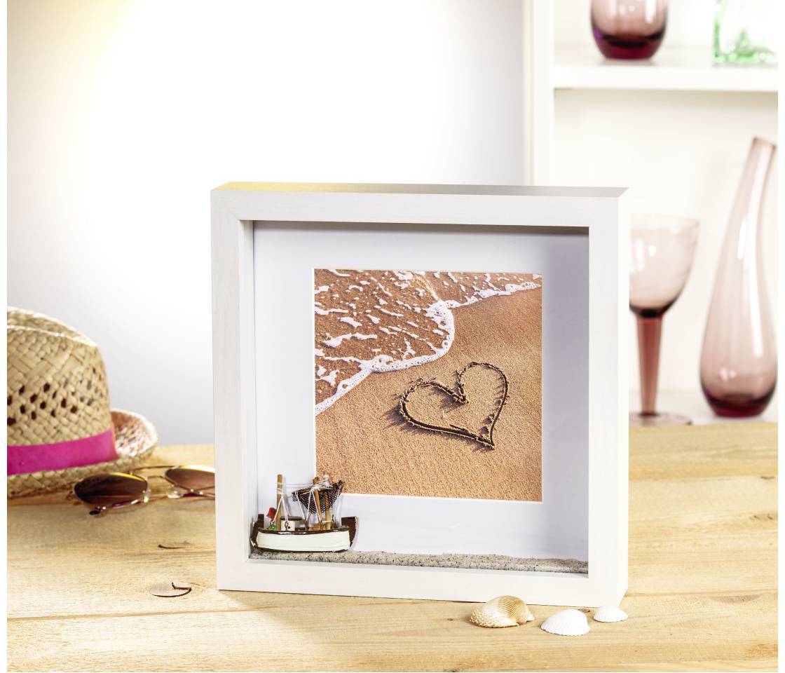 Ein eingerahmtes Bild mit einem Herz im Sand vor einer Welle. Im Vordergrund liegen ein Hut, Sonnenbrille und Muscheln auf einem Tisch.