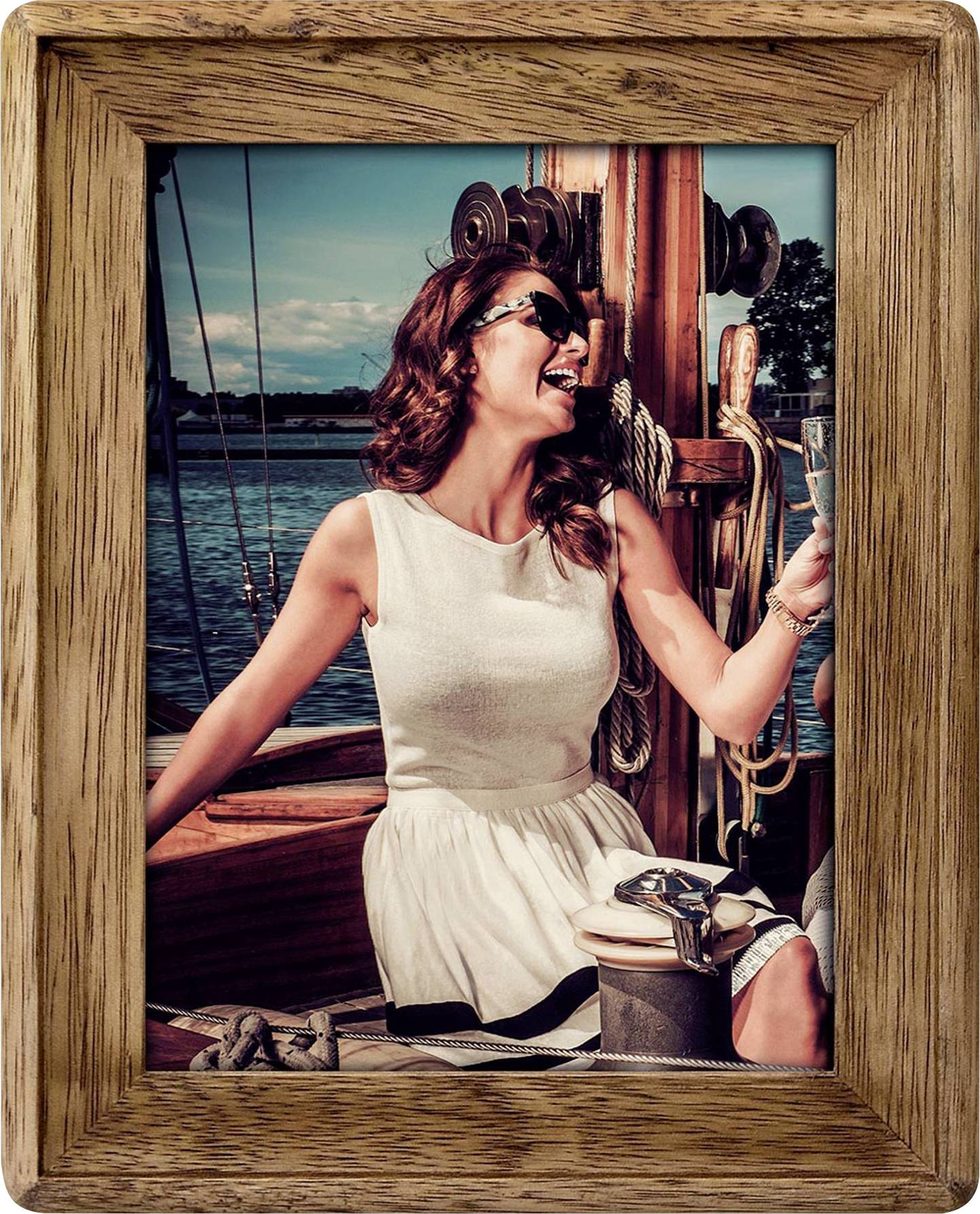 Frau in weißem Kleid sitzt fröhlich auf einem Boot, hält ein Glas Sekt. Im Hintergrund sieht man Wasser und blauen Himmel.