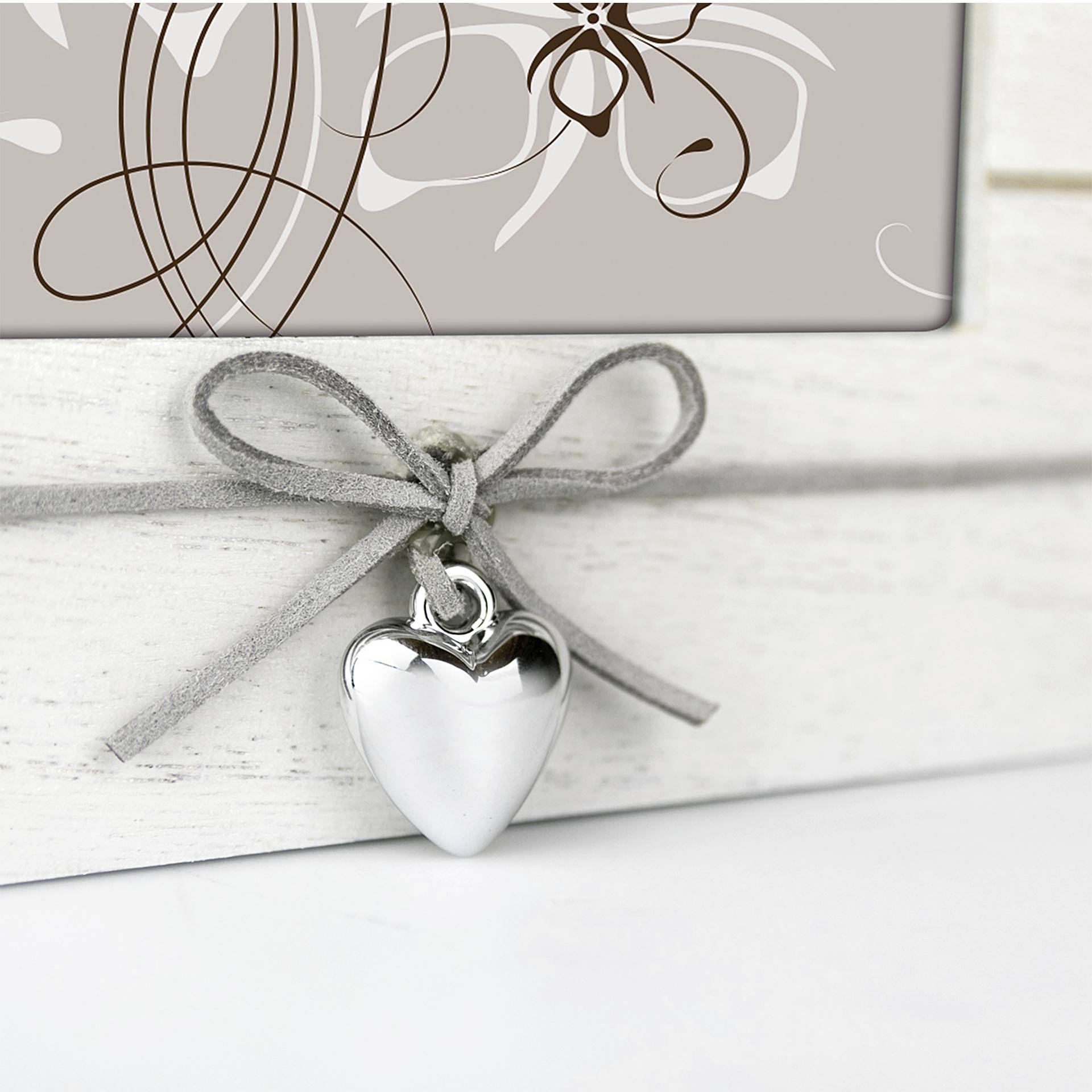 Ein silbernes Herz-Anhänger hängt an einer grauen Schleife vor einem Hintergrund mit abstrakten weißen Blumenmustern auf beigem Grund.