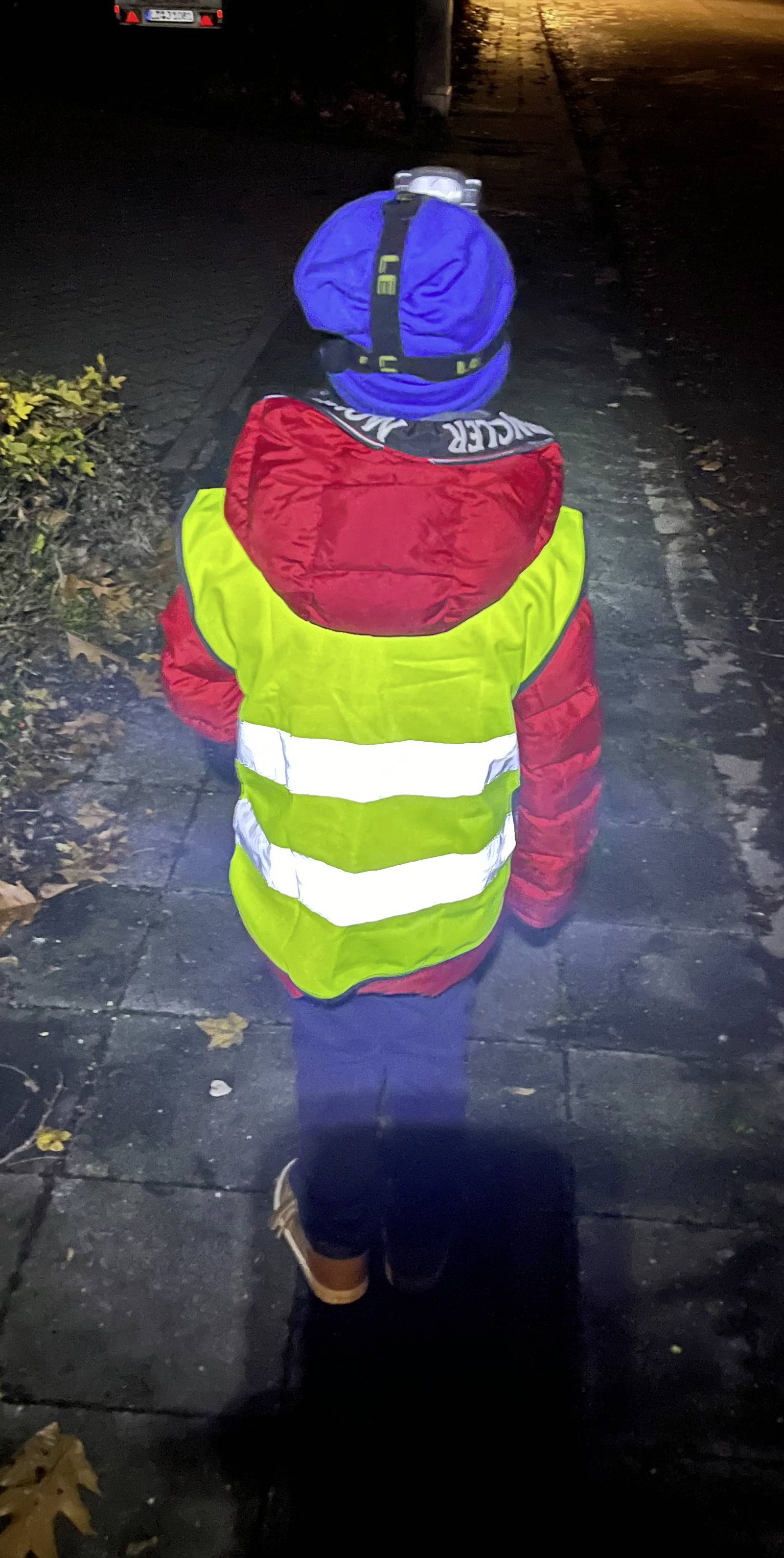 Ein Kind mit blauer Mütze und roter Jacke trägt eine reflektierende gelbe Weste und läuft bei Nacht auf einem Bürgersteig.