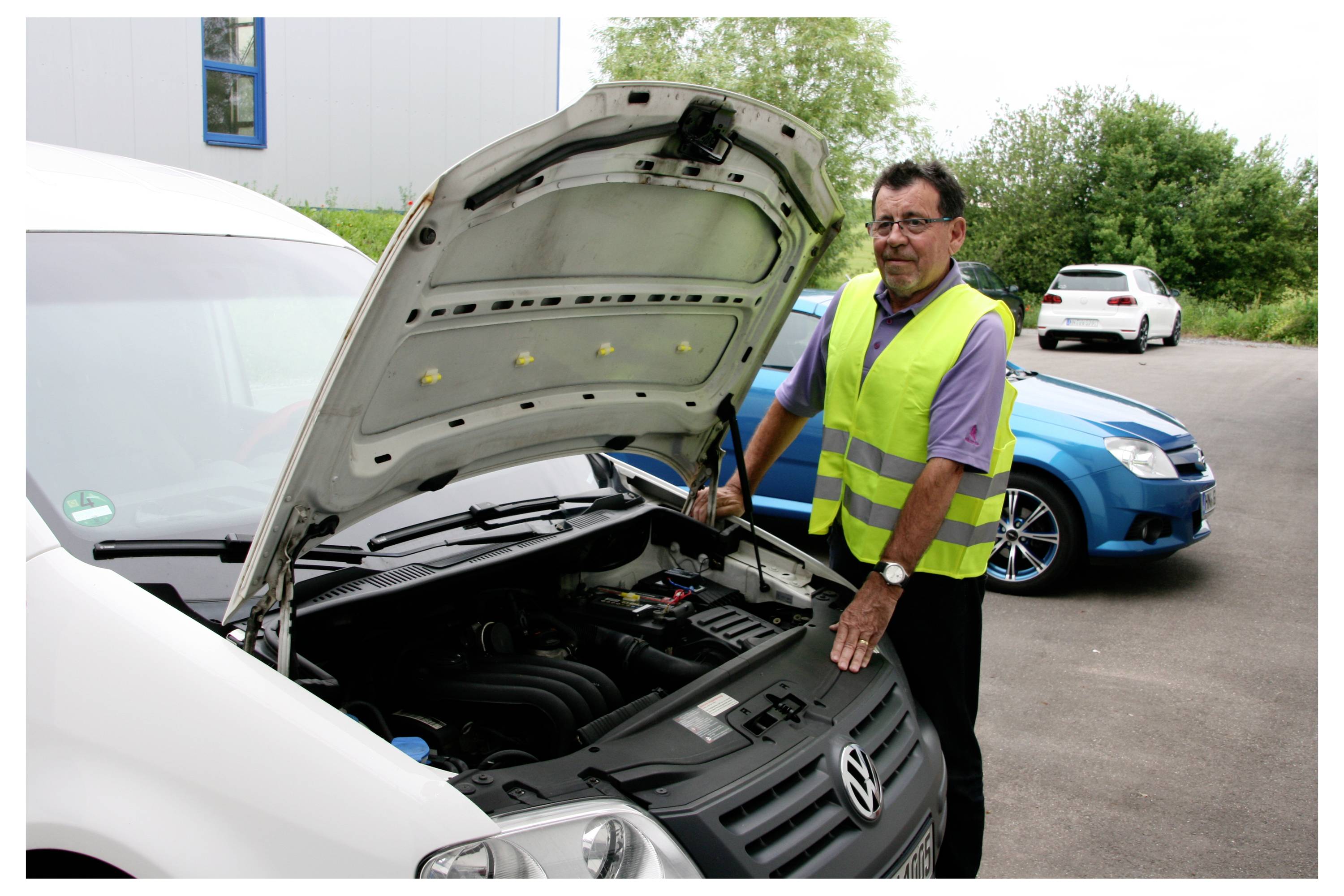 Eine Person in einer Warnweste inspiziert den Motor eines weißen Lieferwagens mit geöffneter Motorhaube. Ein blaues Auto ist in der Nähe geparkt.
