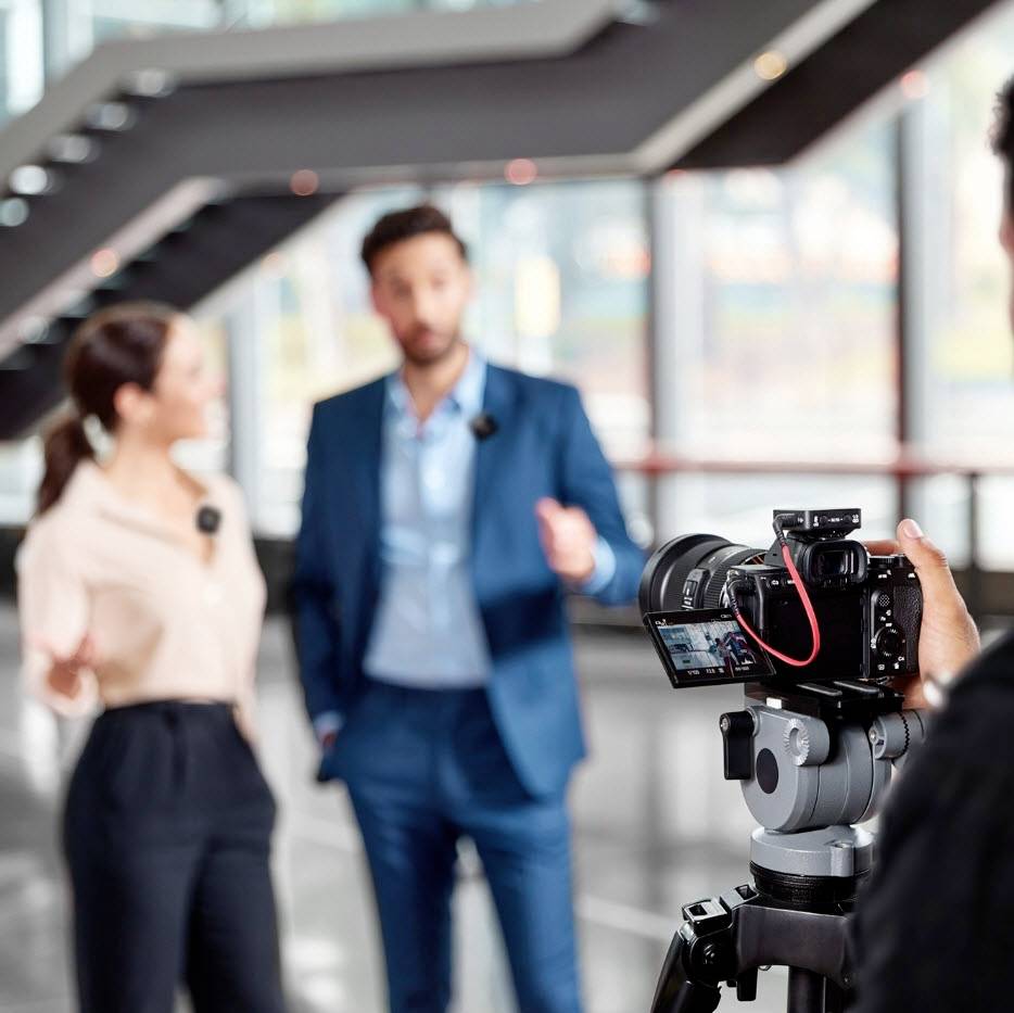 Eine Frau und ein Mann stehen vor einer Kamera in einem modernen Gebäude, offenbar in einer Unterhaltung oder bei einem Interview.