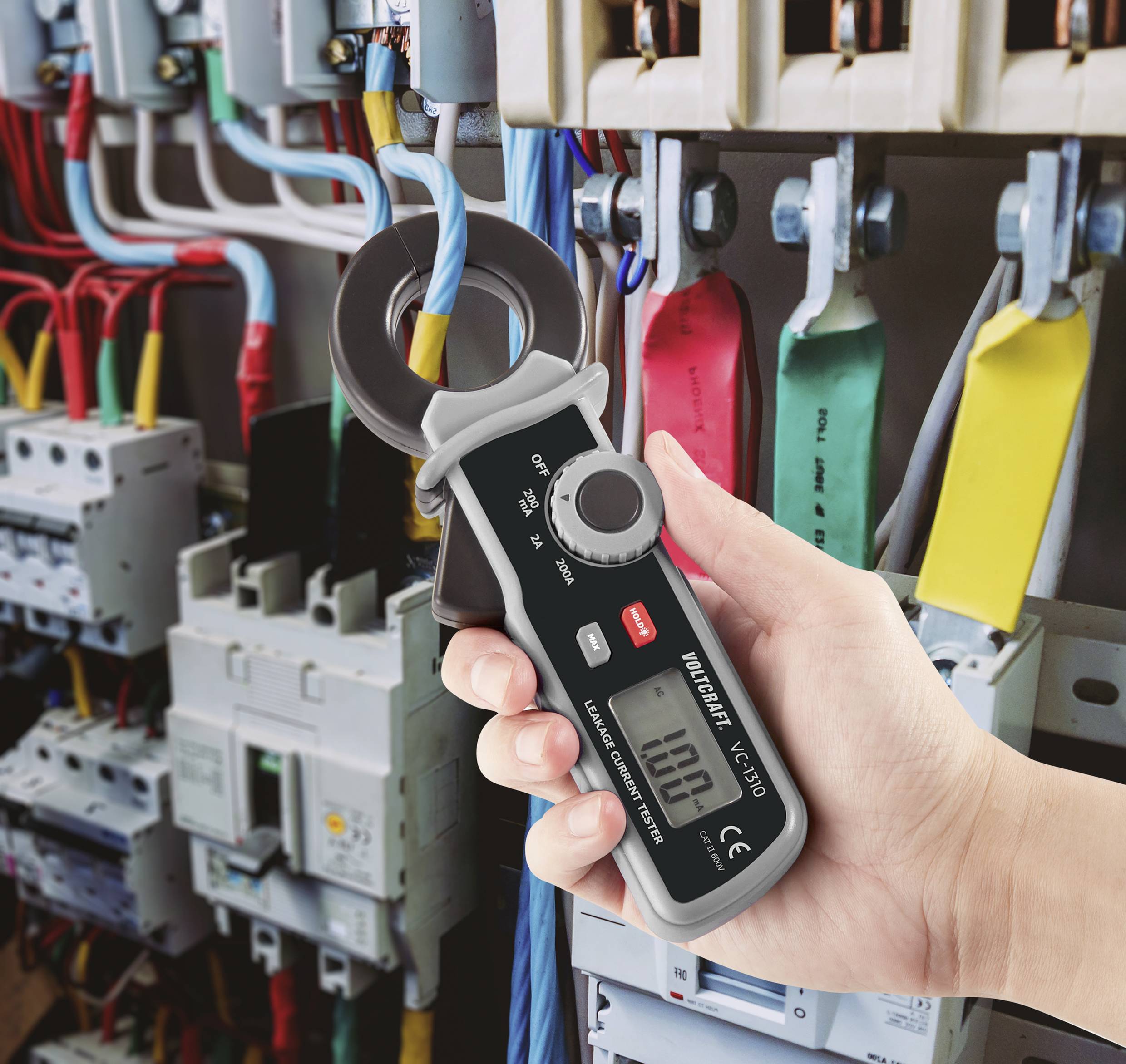 Eine Hand hält ein digitales Multimeter mit Stromzange vor einem offenen Schaltschrank mit farbigen Kabeln und Sicherungen.