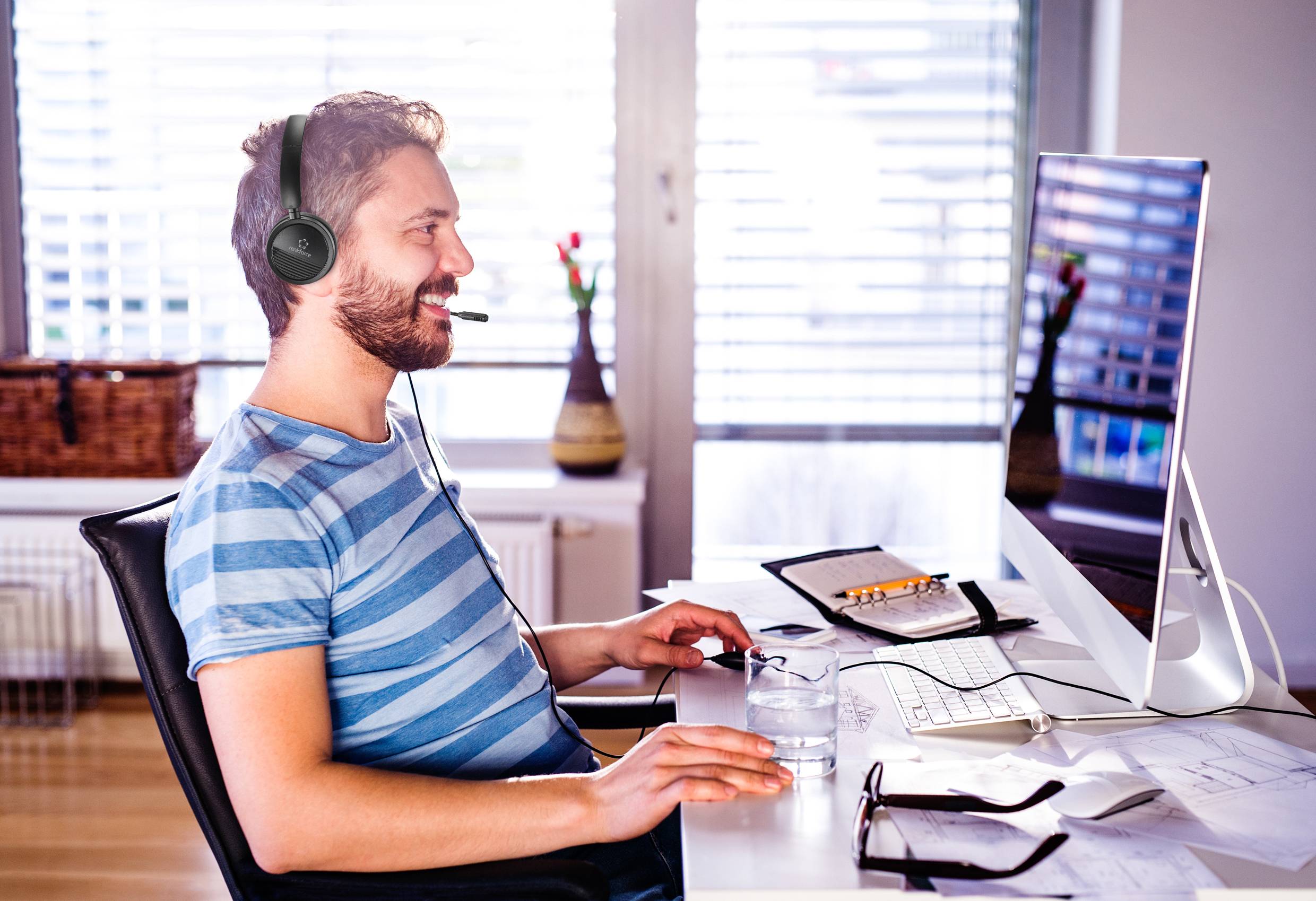 Ein Mann mit Headset sitzt lächelnd am Schreibtisch vor einem Computer. Neben ihm liegen ein Glas Wasser, ein Notizblock und Stifte.