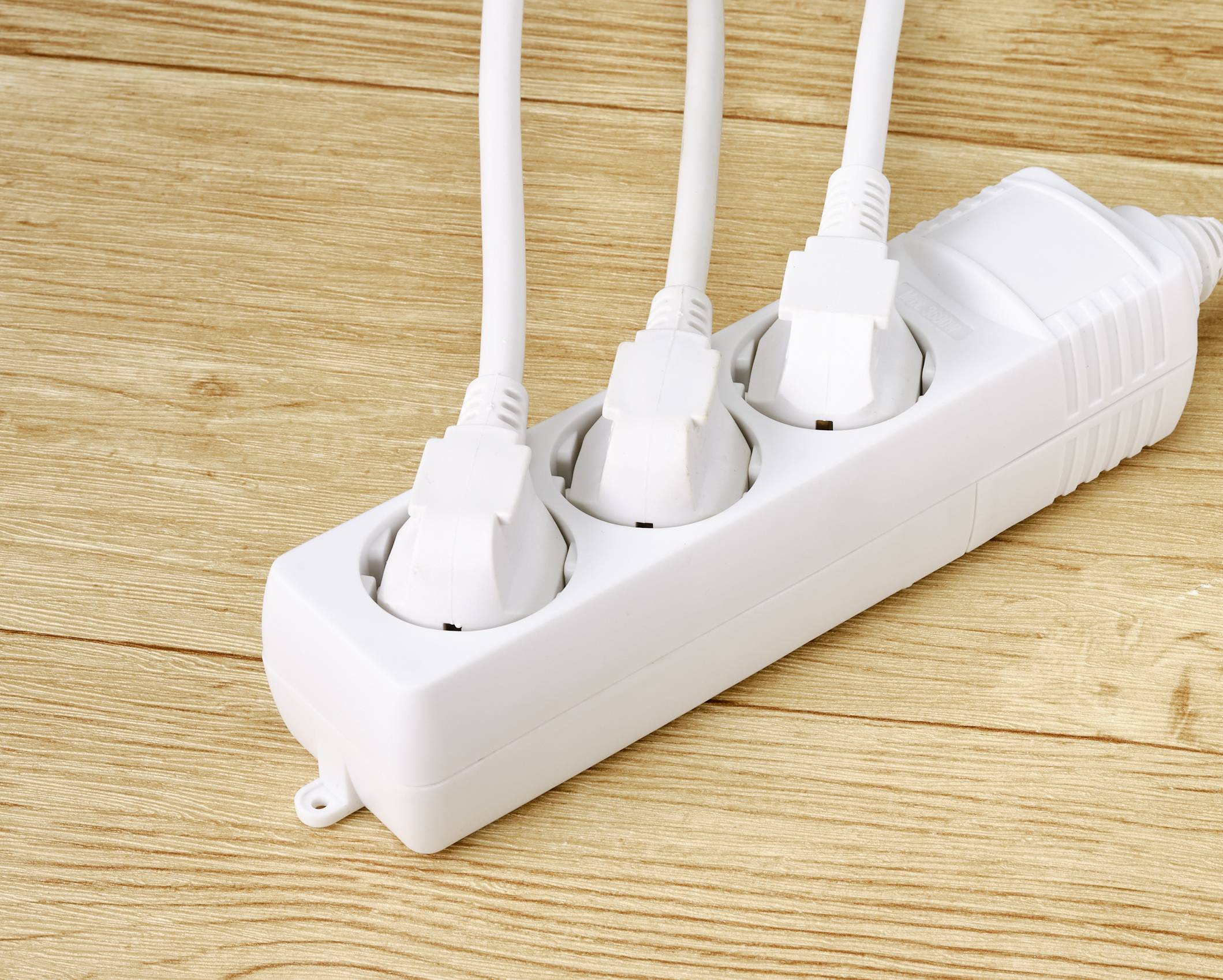 Drei weiße Stecker sind in eine weiße Mehrfachsteckdose auf einem Holzboden eingesteckt.