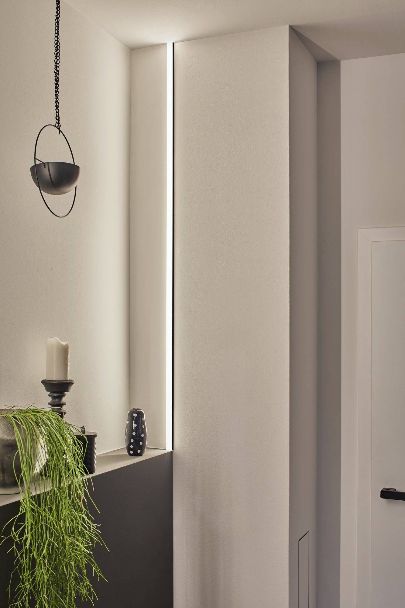 Ein minimalistisches, modernes Interieur mit schmalem Leuchtstreifen an der Wand, schlichtem Kerzenhalter, dekorativer Vase und einer Hängepflanze.