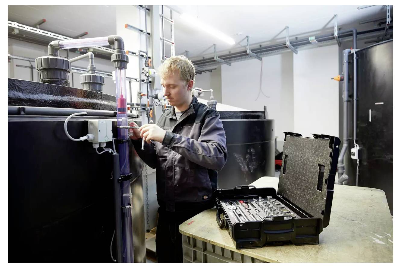 Eine Person in einer Werkstatt stellt Ausrüstungen an einem Rohr neben großen Tanks ein. Ein offener Werkzeugkasten befindet sich in der Nähe auf einer Werkbank.