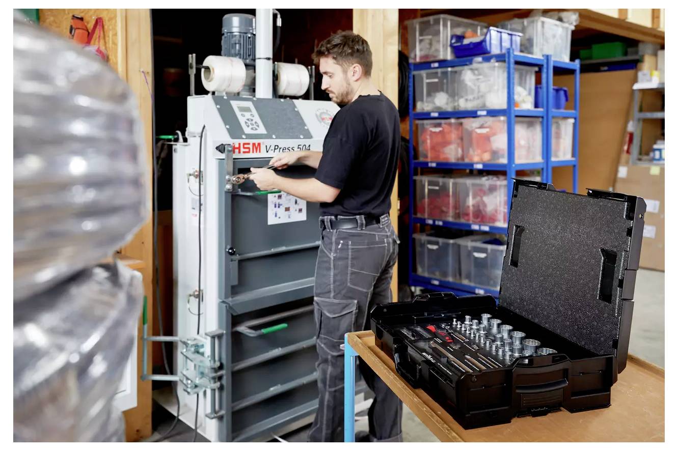 Eine Person bedient eine HSM-Präsmaschine in einer Werkstatt, mit organisierten Regalen und einem Werkzeugkasten in der Nähe, was auf konzentrierte Arbeitsaktivität hinweist.