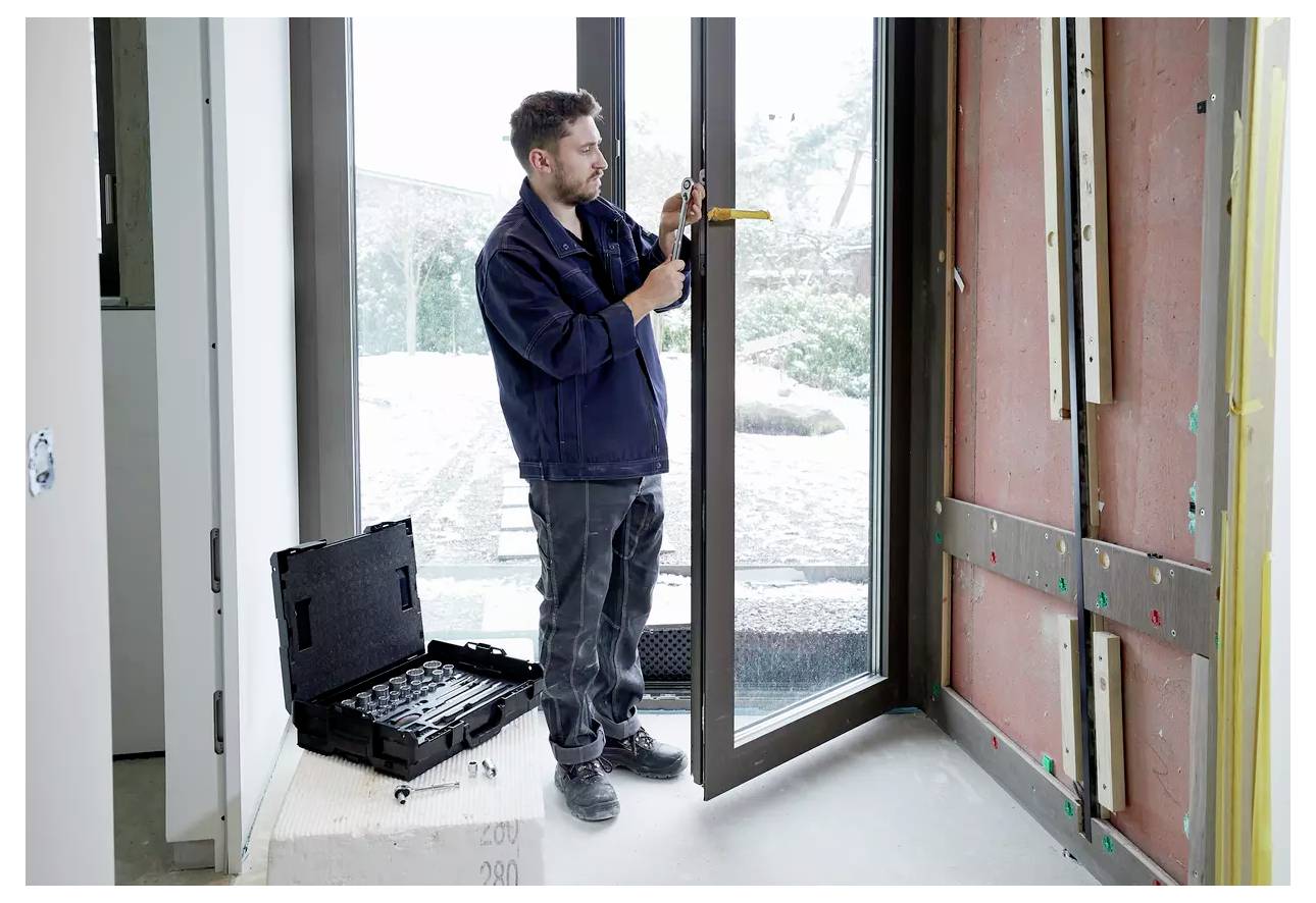 Ein Mann installiert eine große Glastür und verwendet dabei ein Werkzeugset, das in der Nähe positioniert ist. Der Raum befindet sich im Umbau, mit freiliegenden Wänden und Fenstern.