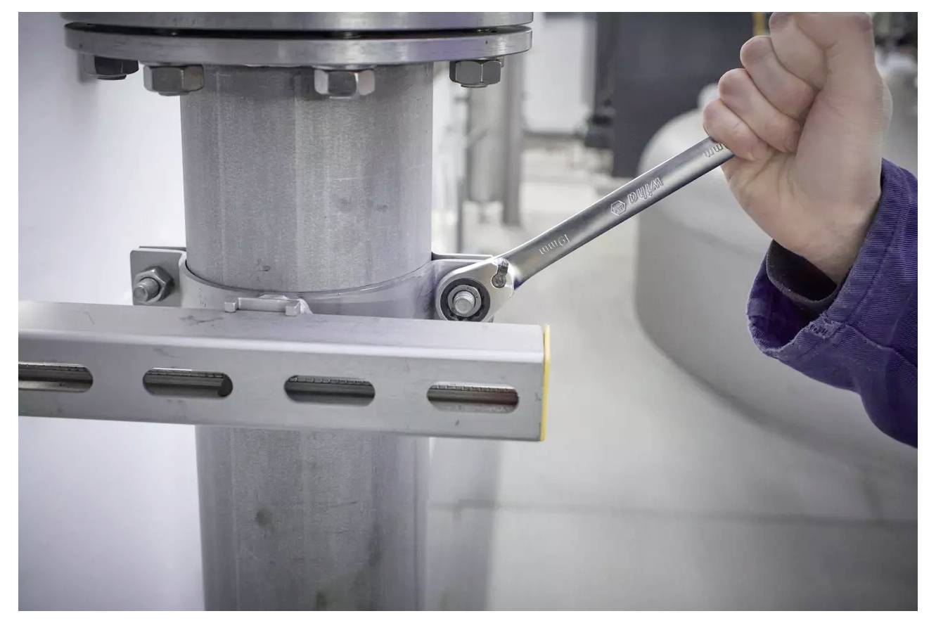 Eine Person verwendet einen Schraubenschlüssel, um eine Schraube an einem Metallpfeiler festzuziehen, was auf Wartungs- oder Montagearbeiten in einer industriellen Umgebung hinweist.