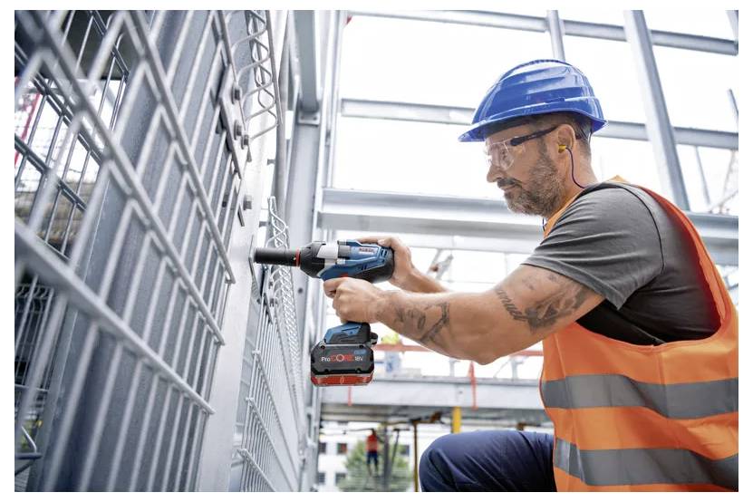 Bauarbeiter in Schutzhelm und Warnweste, der einen Elektroschrauber verwendet, um einen Metallzaun oder ein Metallpaneel an einem Gebäuderahmen zu befestigen.