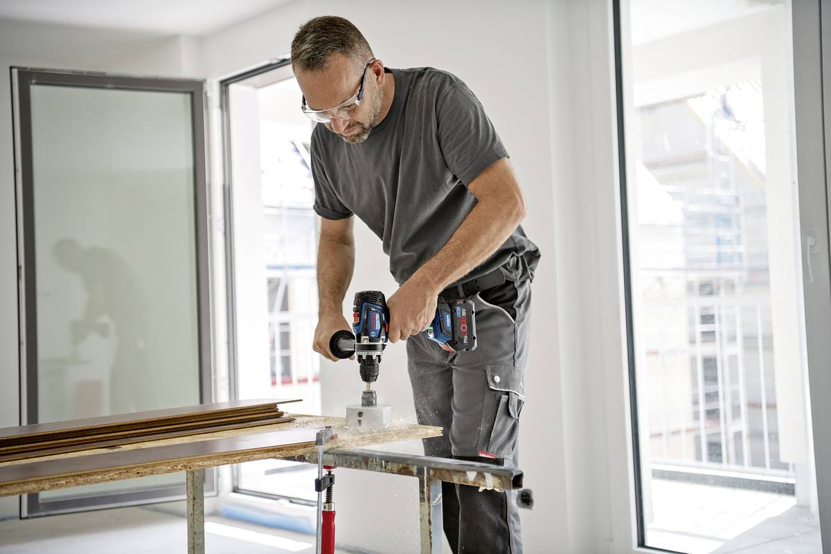 Ein Mann arbeitet mit einer Bohrmaschine an einem Tisch in einem hellen Raum mit großen Fenstern. Schutzbrille und professionelle Kleidung.
