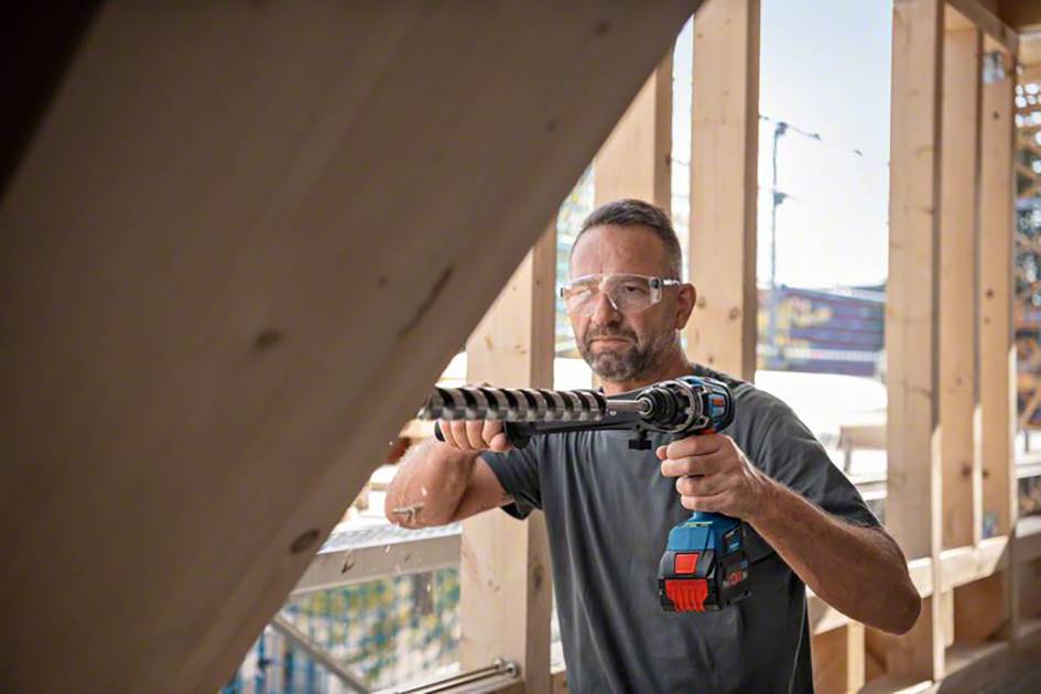 Ein Mann mit Schutzbrille arbeitet mit einer Bohrmaschine an einem Holzrahmen auf einer Baustelle. Sonnenlicht fällt durch die Struktur.