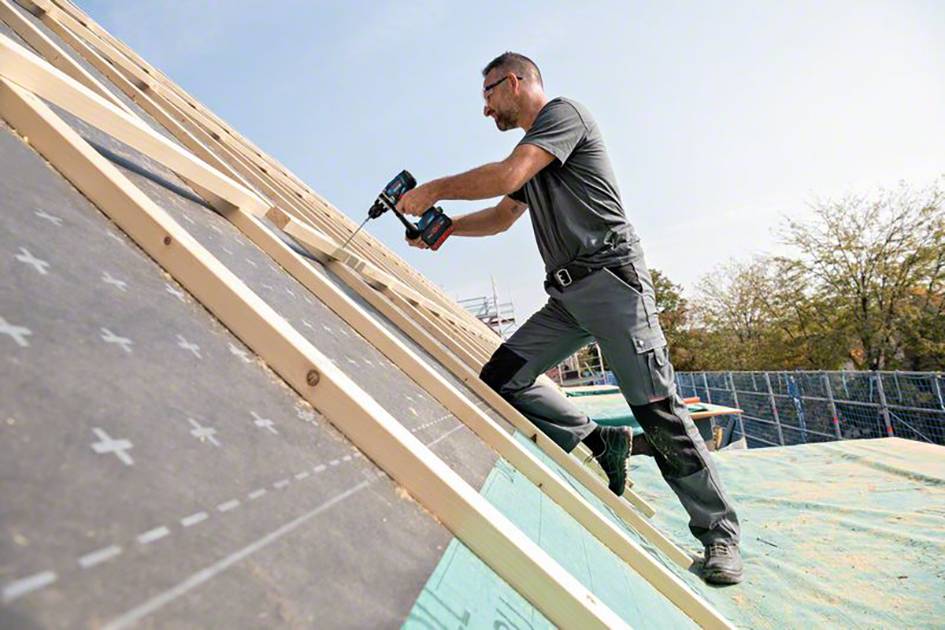 Ein Mann arbeitet auf einem Dach, bohrt Holzlatten mit einem Elektrowerkzeug. Er trägt Schutzkleidung und die Umgebung zeigt ein Baugerüst.