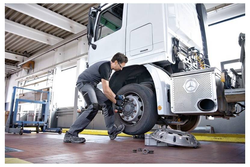 Ein Mechaniker wechselt das Rad eines großen Lastkraftwagens in einer Autowerkstatt-Bucht, mit dem angehobenen Reifen und Werkzeugen auf dem Boden.