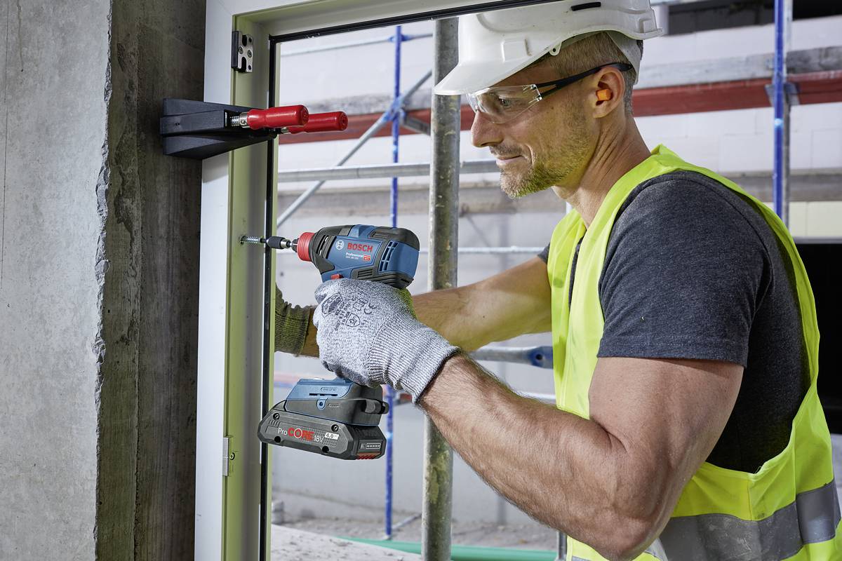 Ein Bauarbeiter in Schutzkleidung montiert ein Fensterrahmen in einer Baustelle mit einer Bohrmaschine. Er trägt Helm und Handschuhe.