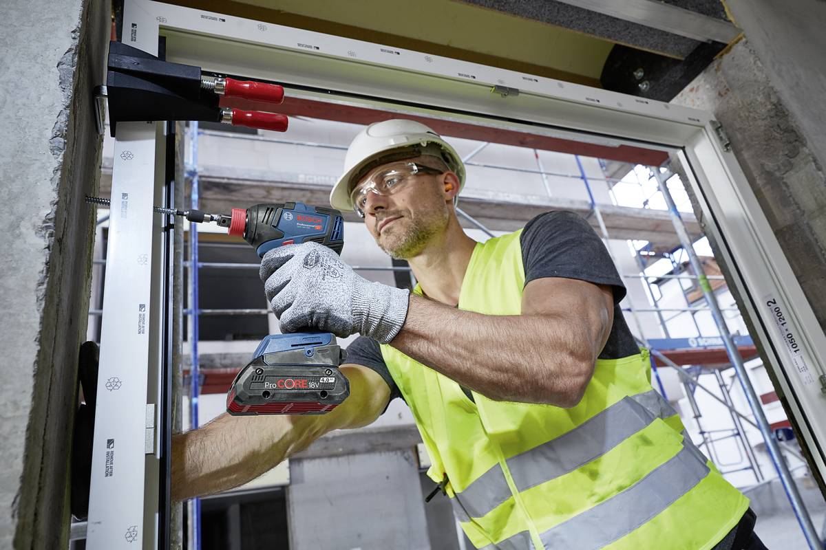Ein Bauarbeiter mit Schutzhelm und Weste benutzt eine Bohrmaschine, um in einem Gebäude an einer Fensteröffnung zu arbeiten. Im Hintergrund ist ein Baugerüst sichtbar.