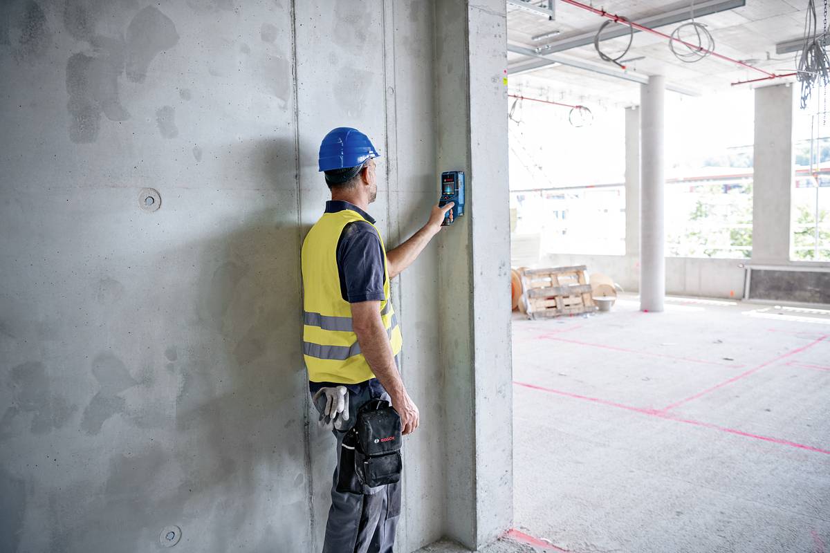 Ein Bauarbeiter mit Schutzhelm und Warnweste misst mit einem Gerät die Wand eines Rohbaus. Umgebung zeigt unvollendeten Bau.
