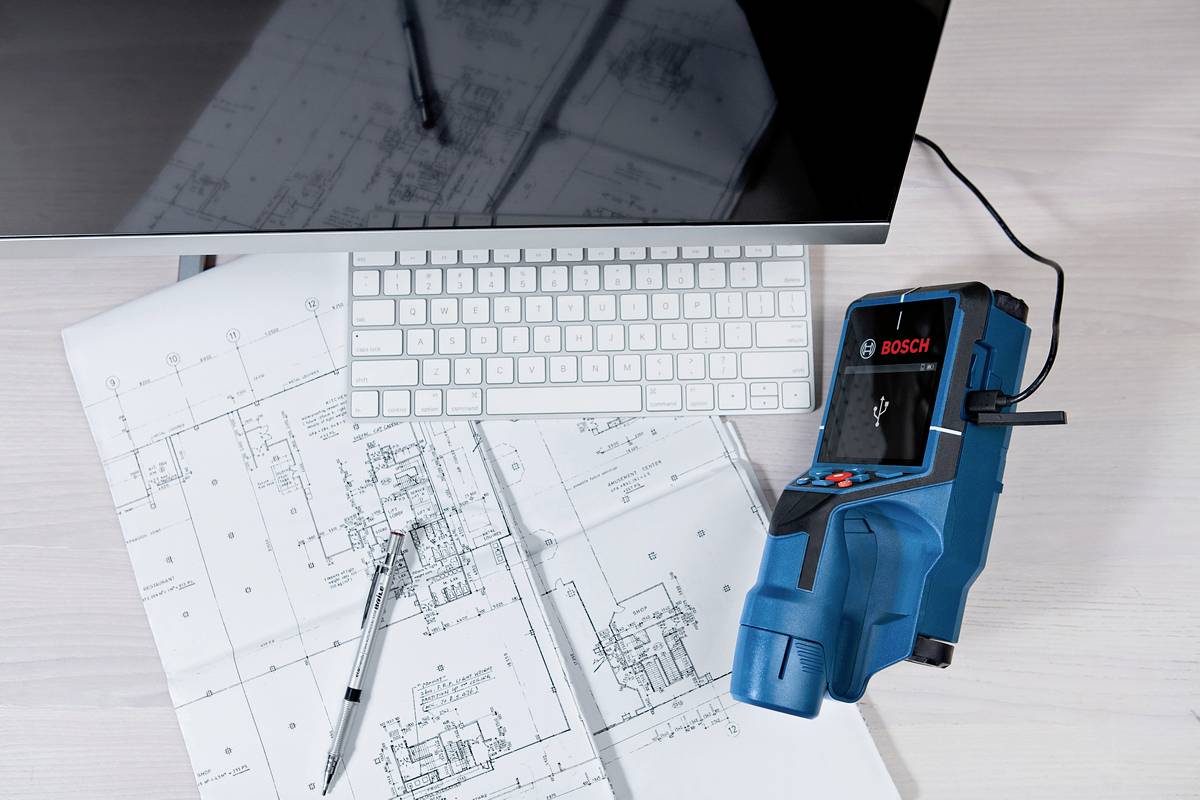Eine Tastatur, ein Computerbildschirm, Baupläne und ein blaues Bosch-Messgerät liegen auf einem Tisch. Ein Stift liegt auf den Plänen.