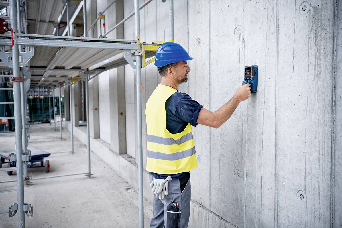 Ein Bauarbeiter in gelber Weste und blauem Helm nutzt ein Messgerät an einer Betonwand auf einer Baustelle mit Gerüsten.