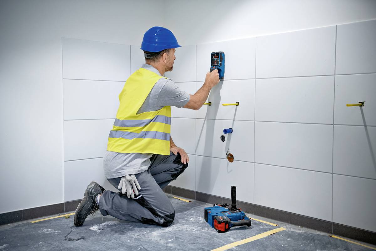 Ein Bauarbeiter kniet am Boden und misst die Feuchtigkeit einer gefliesten Wand mit einem elektronischen Gerät. Er trägt gelbe Sicherheitsweste und blauen Helm.