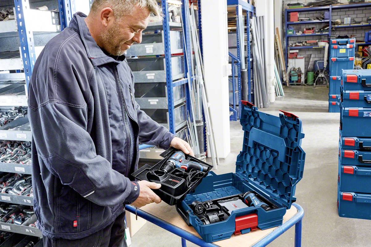 Ein Mann im Lagerraum öffnet einen Koffer mit Elektrowerkzeugen, darunter eine Bohrmaschine. Regale mit Werkzeugen im Hintergrund.