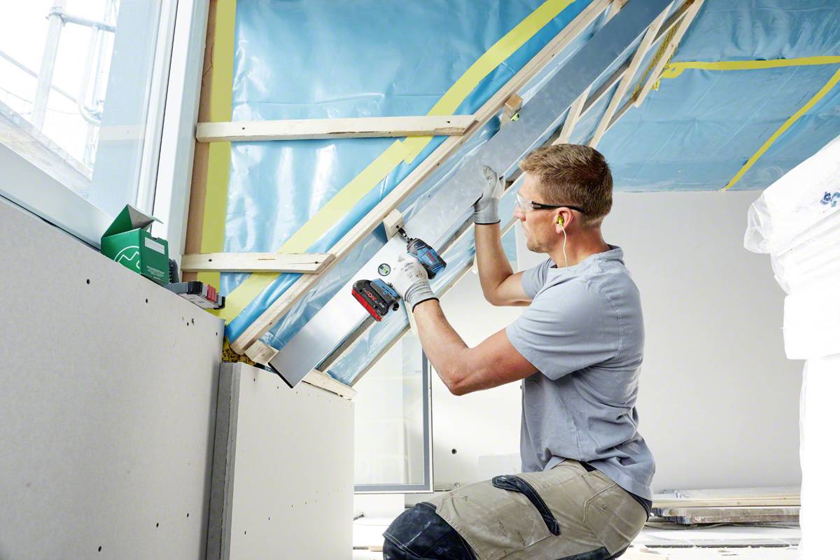 Ein Arbeiter montiert mit einem Akkubohrer eine Dachverkleidung. Er trägt Schutzbrille und Ohrstöpsel. Im Hintergrund sind Dämmmaterialien sichtbar.